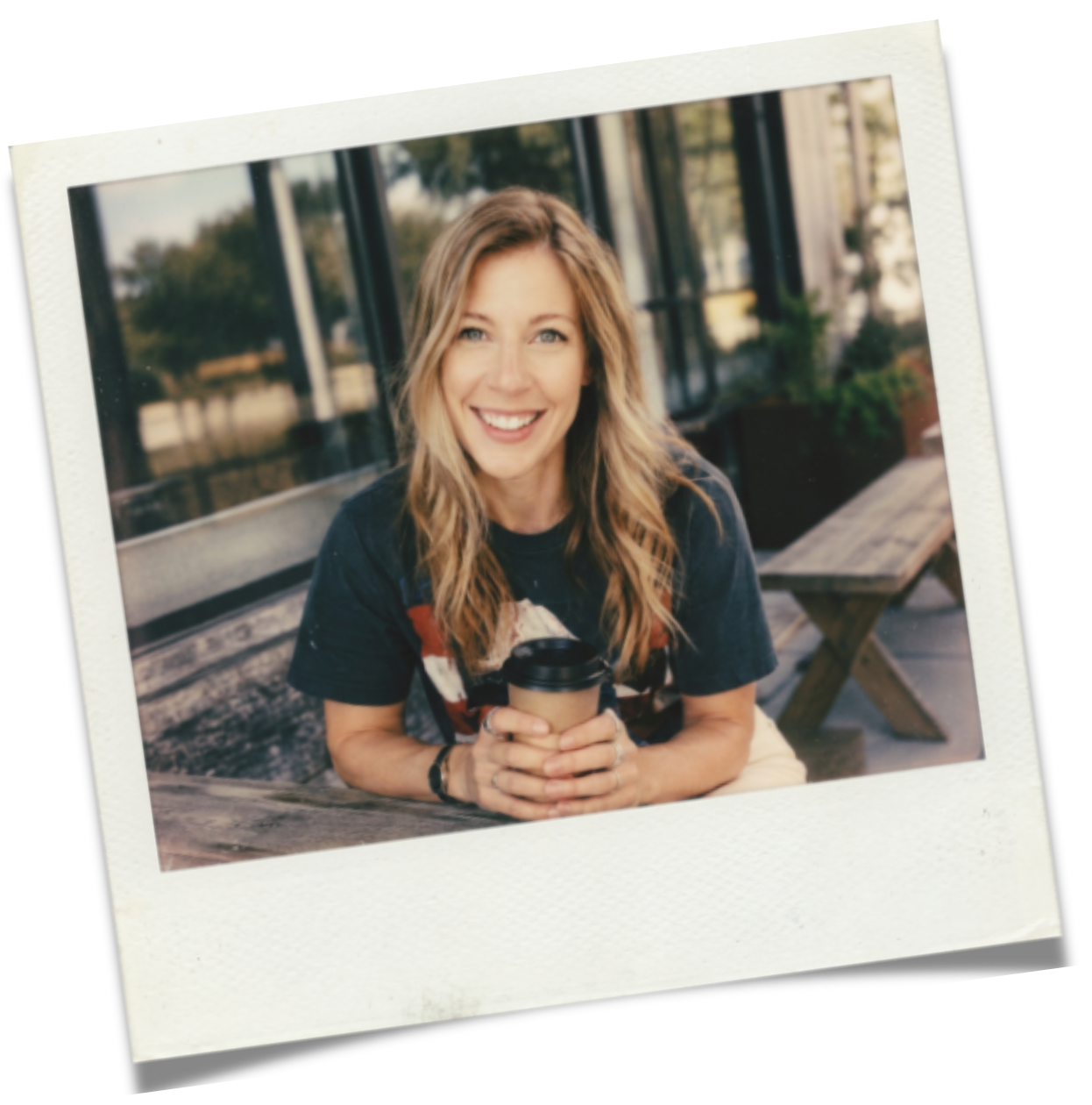 A smiling woman with wavy blonde hair sitting outdoors at a wooden table, holding a coffee cup, with a blurred outdoor background.