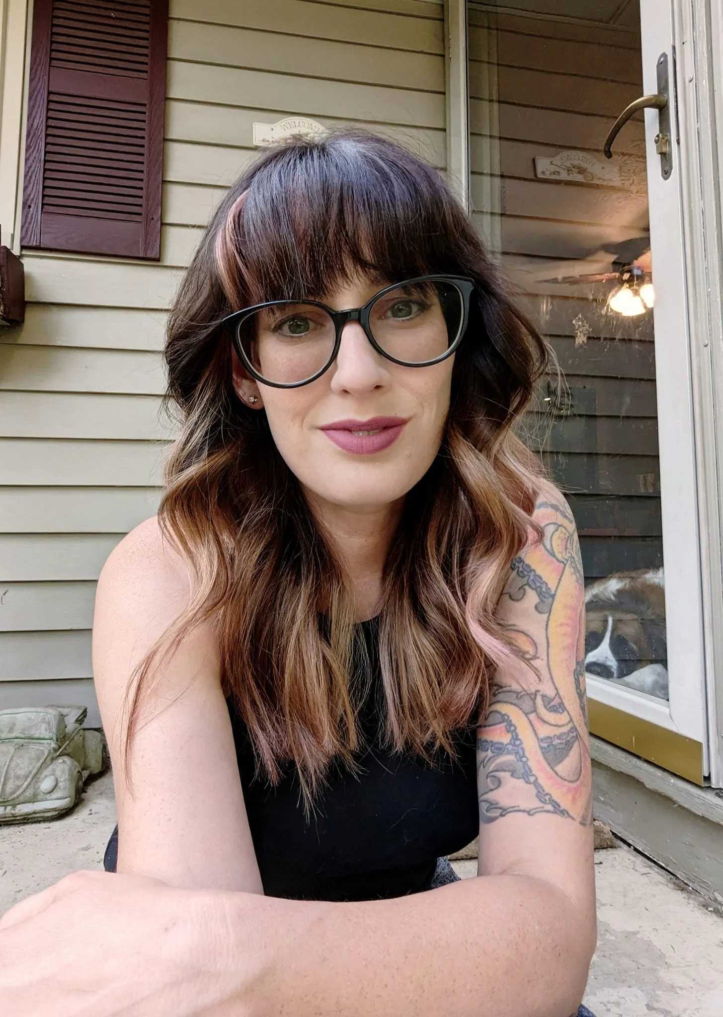 A woman with dark hair and glasses sitting outside on a concrete surface near a house with tan siding and a window with purple shutters, a dog visible through a glass door behind her.