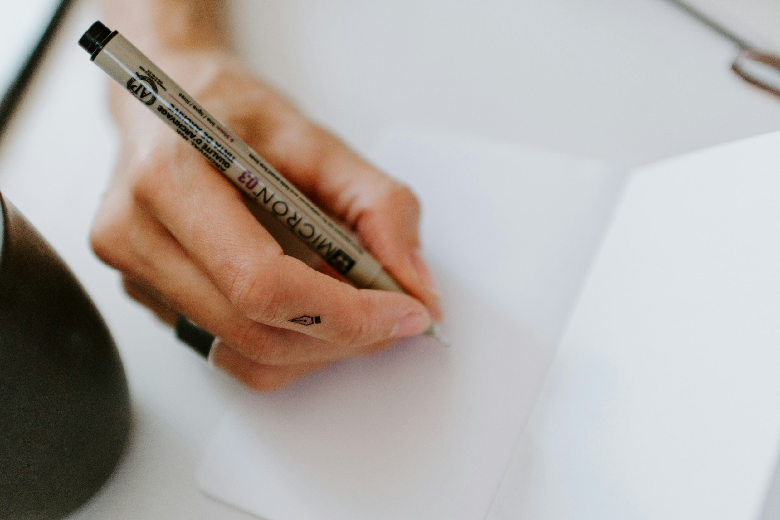 Close-up of a person’s hand holding a black and white permanent marker with the cap on, writing on a white surface, with black object partially visible in the foreground.