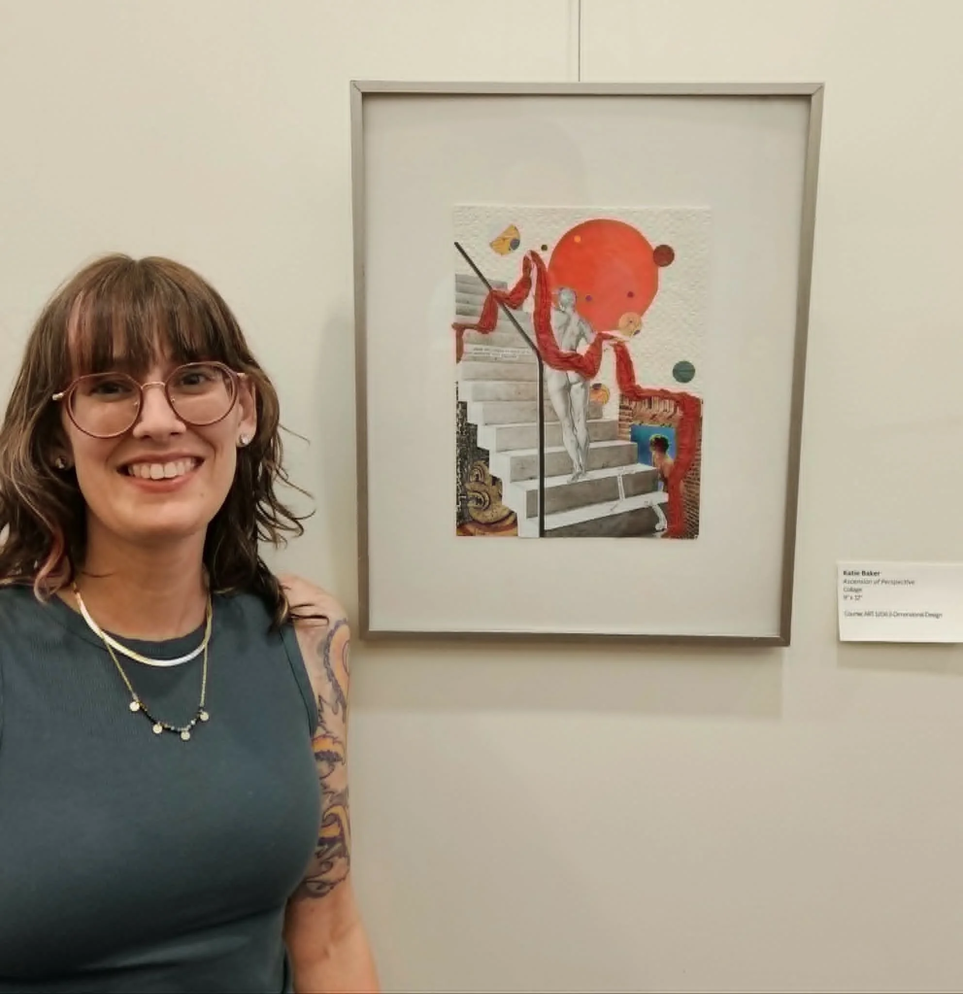 Part of a woman with glasses, curly brown hair, and colorful tattoos on her left arm smiling in front of an abstract colorful art piece on the wall.