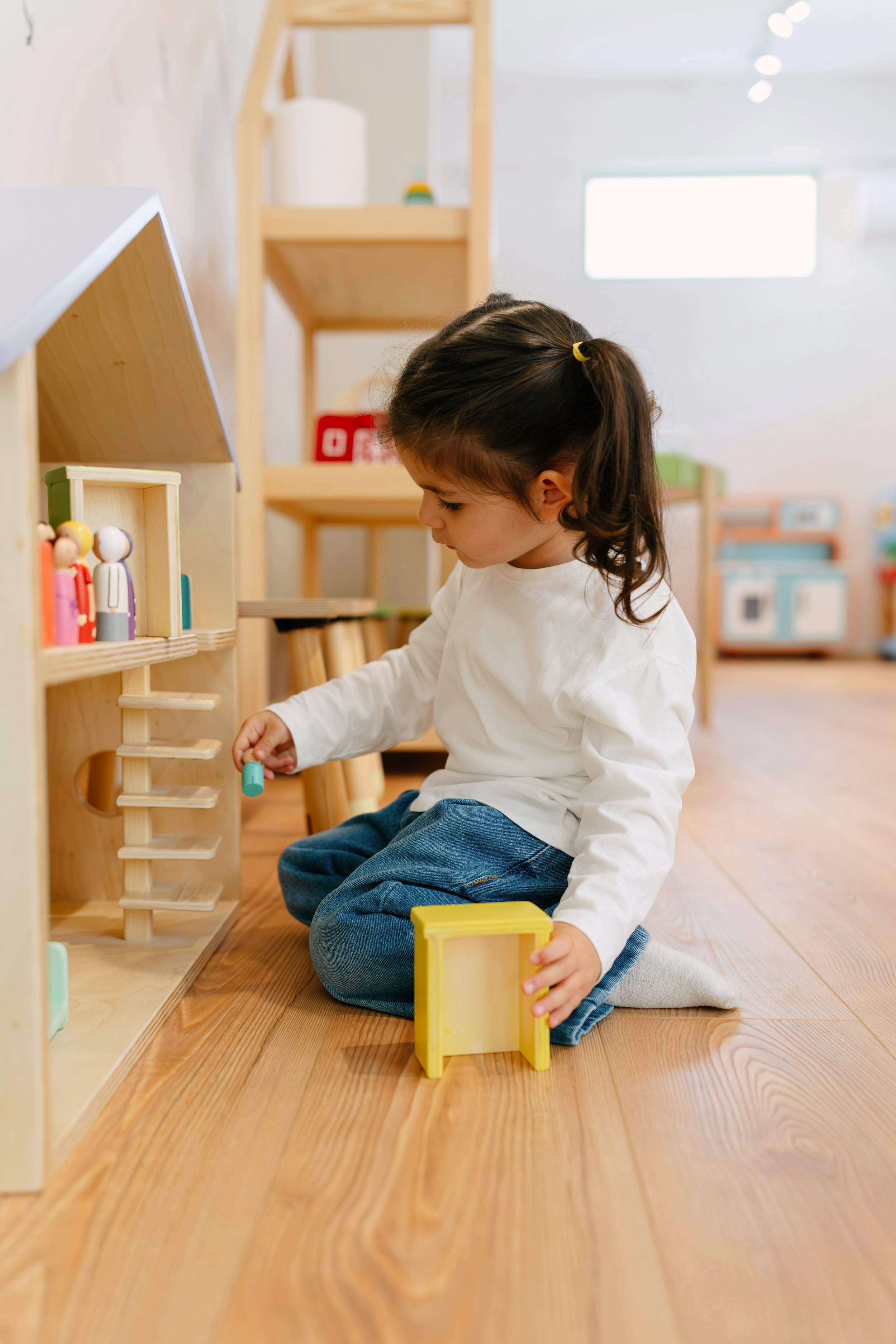 Child kneeling on wooden floor, playing with colorful toy blocks in a bright room with wooden furniture and toys.