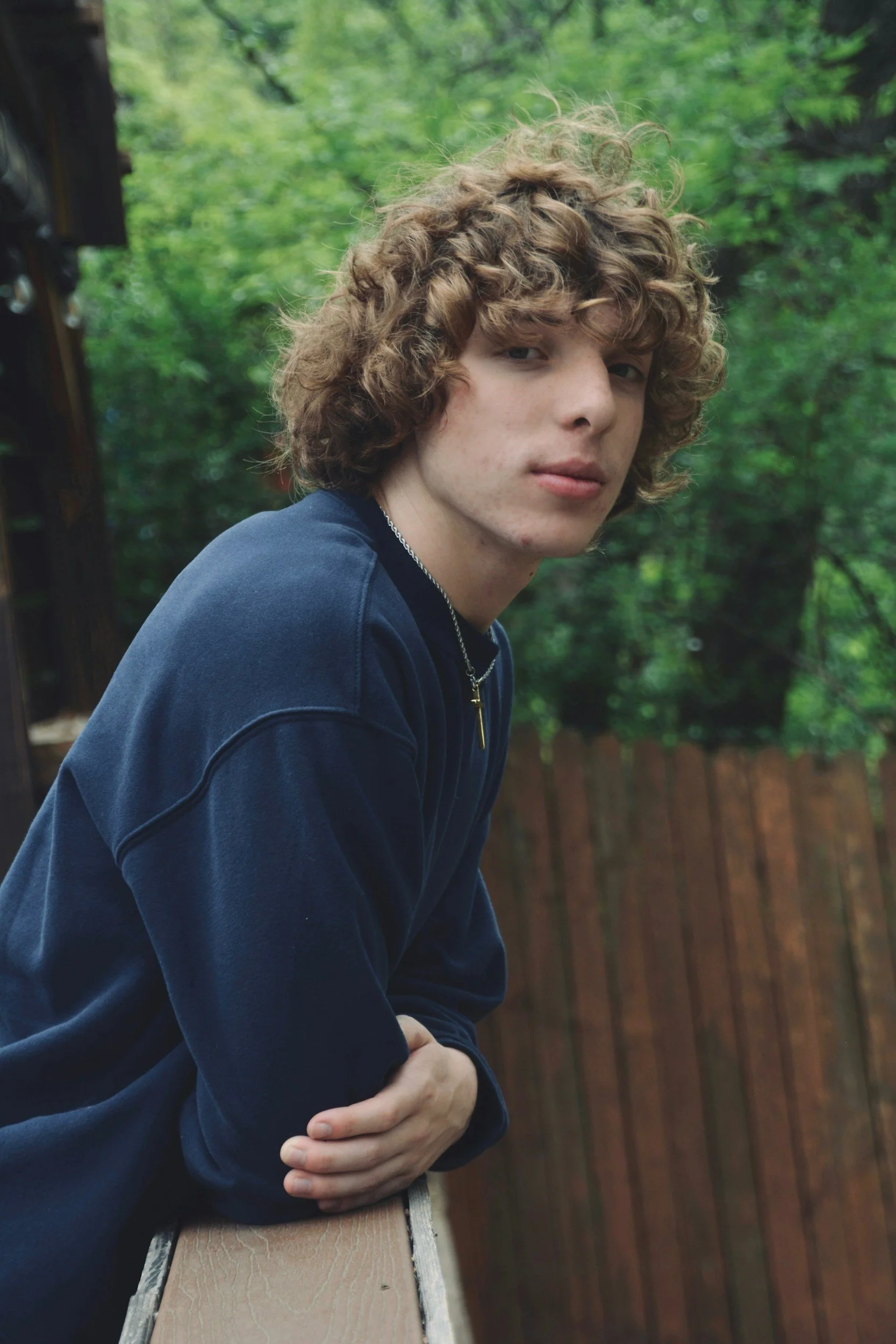 A young man with curly brown hair and a pale complexion, wearing a dark blue sweatshirt, standing outdoors with a blurred green and brown background, arms crossed.
