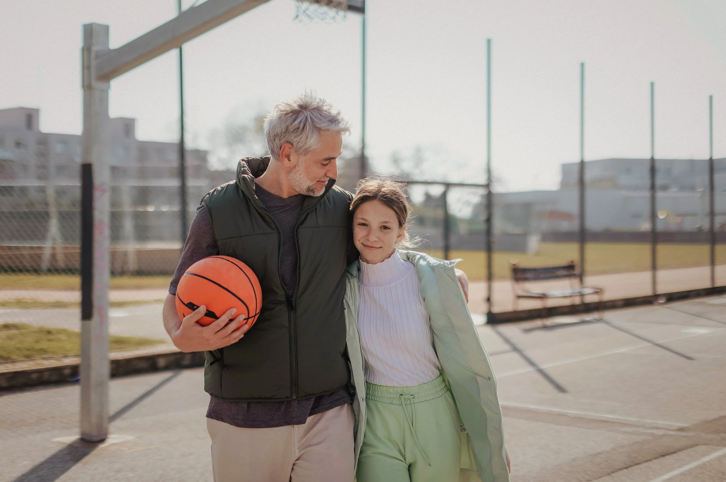 A man with gray hair and beard holding an orange basketball, walking beside a young girl with long hair, both smiling and walking together near a basketball court on a clear day.