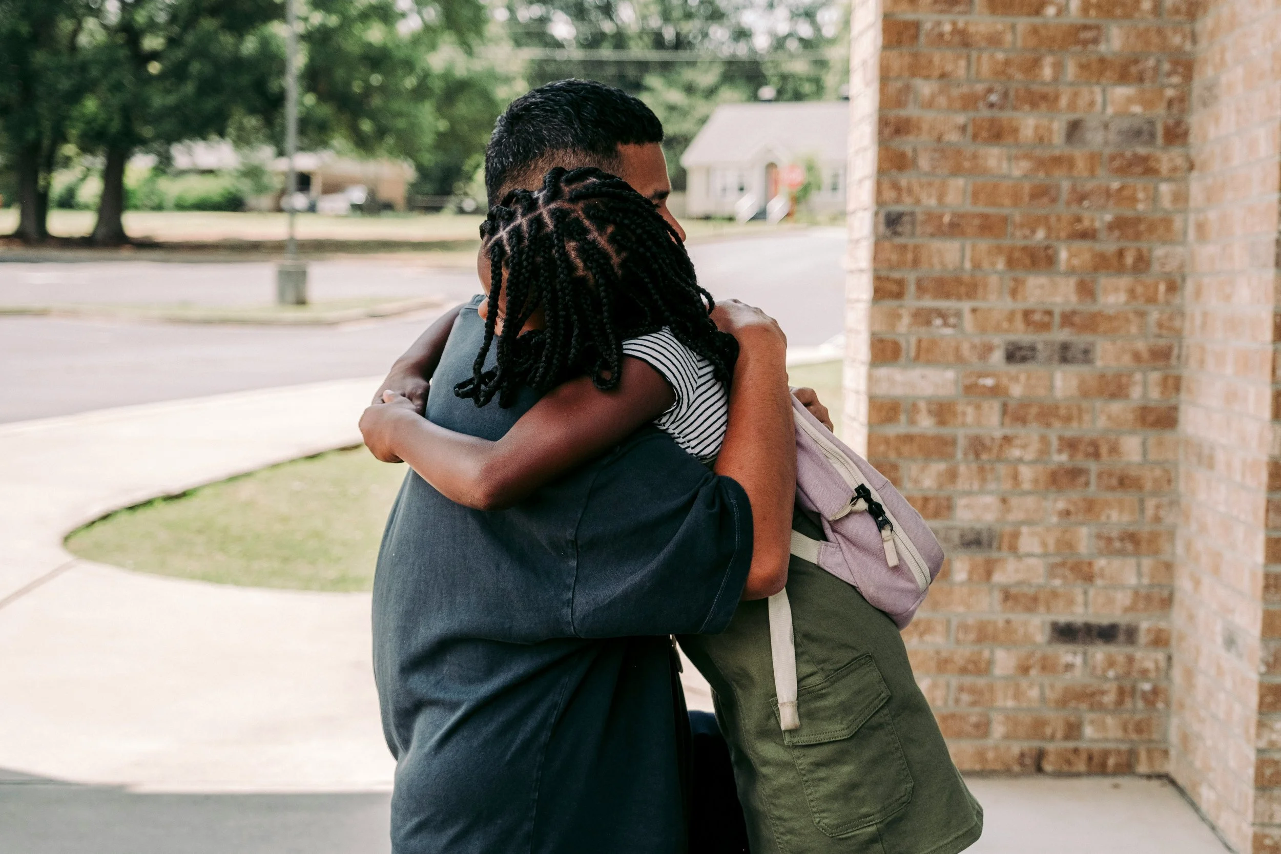 Two people hugging outside near a brick wall with trees in the background.