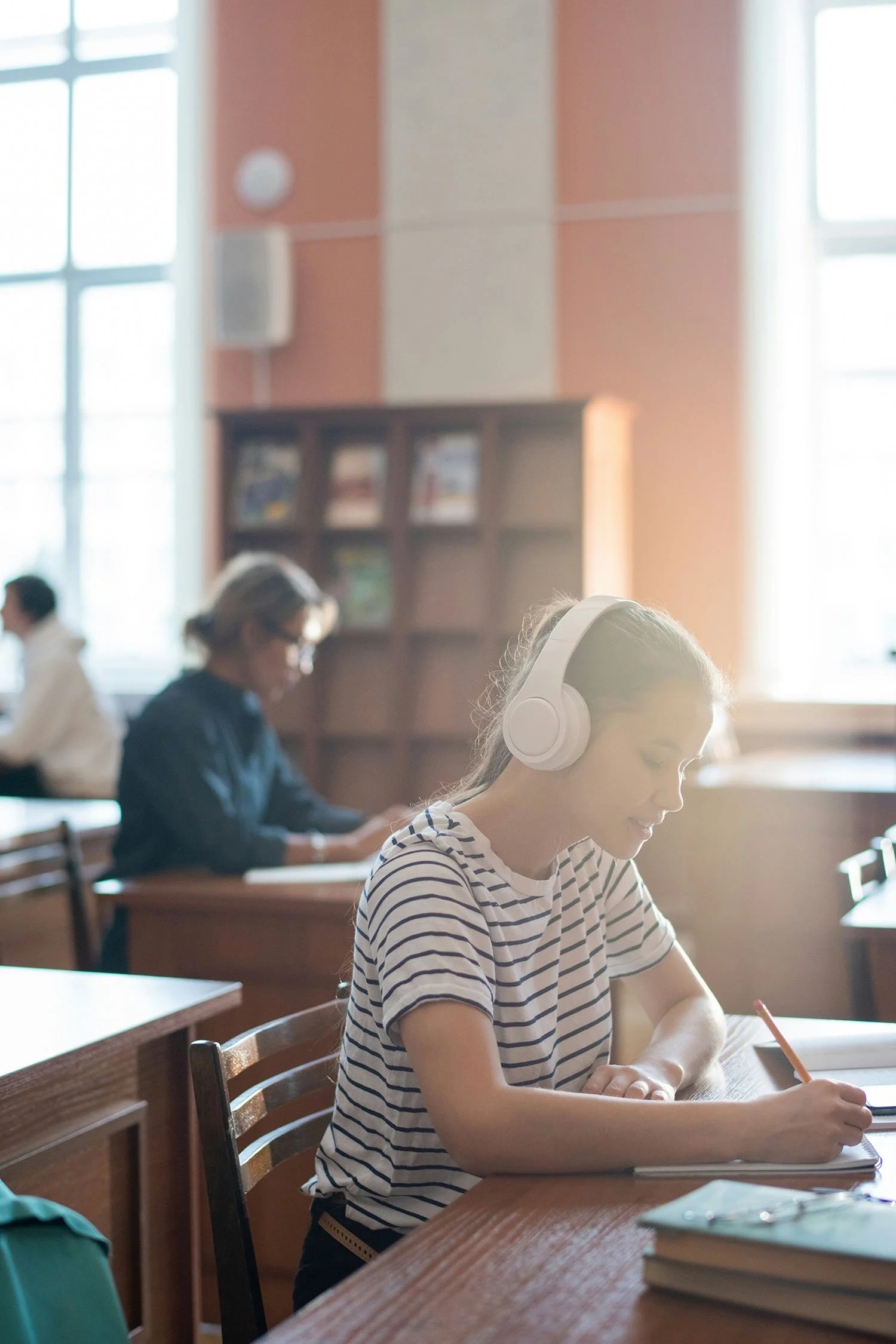 A young girl wearing a striped shirt and white headphones sitting at a wooden desk in a classroom, writing in a notebook. Other students are in the background, and the classroom has large windows with sunlight coming through.