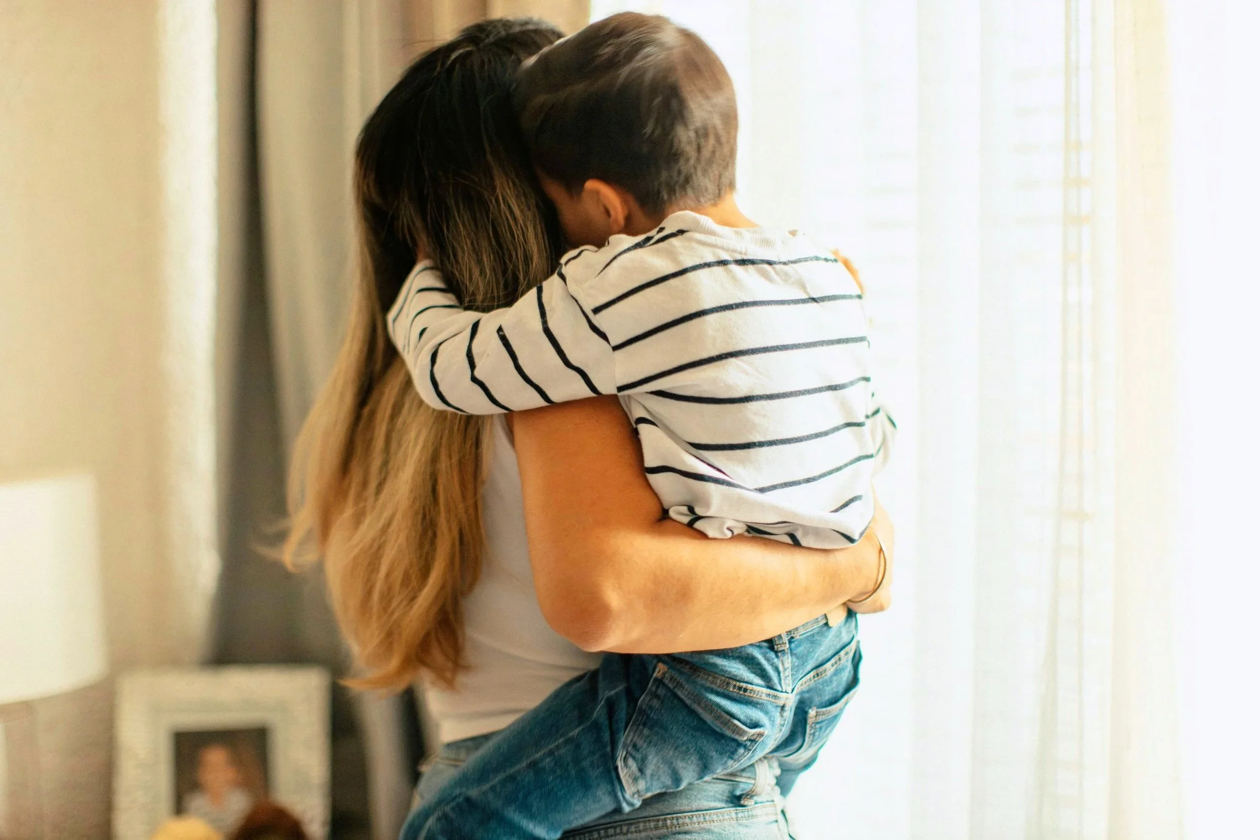 A woman holding a young child in her arms, with the child embracing her and their faces close together, indoors near a window with sheer curtains.