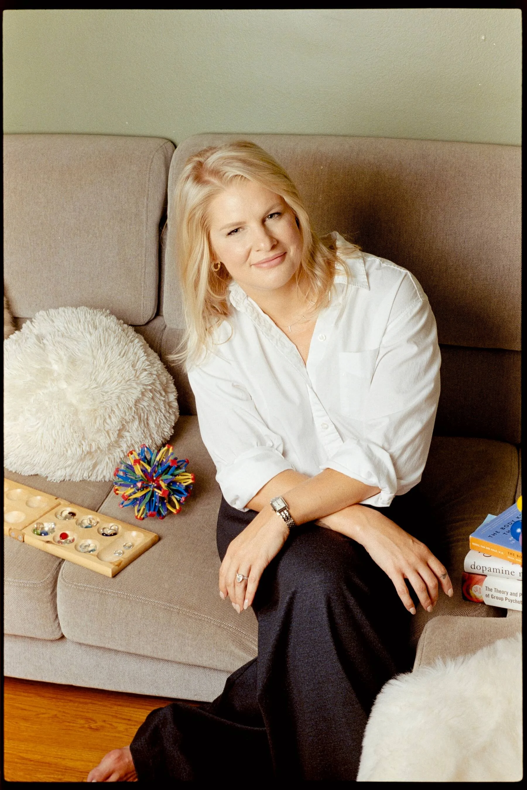 A woman with blonde hair sitting on a beige couch, smiling at the camera. She is wearing a white shirt and a watch. On the couch beside her are a fluffy beige pillow, a wooden board with colorful marbles, and a pile of books including titles related to psychology and dopamine.