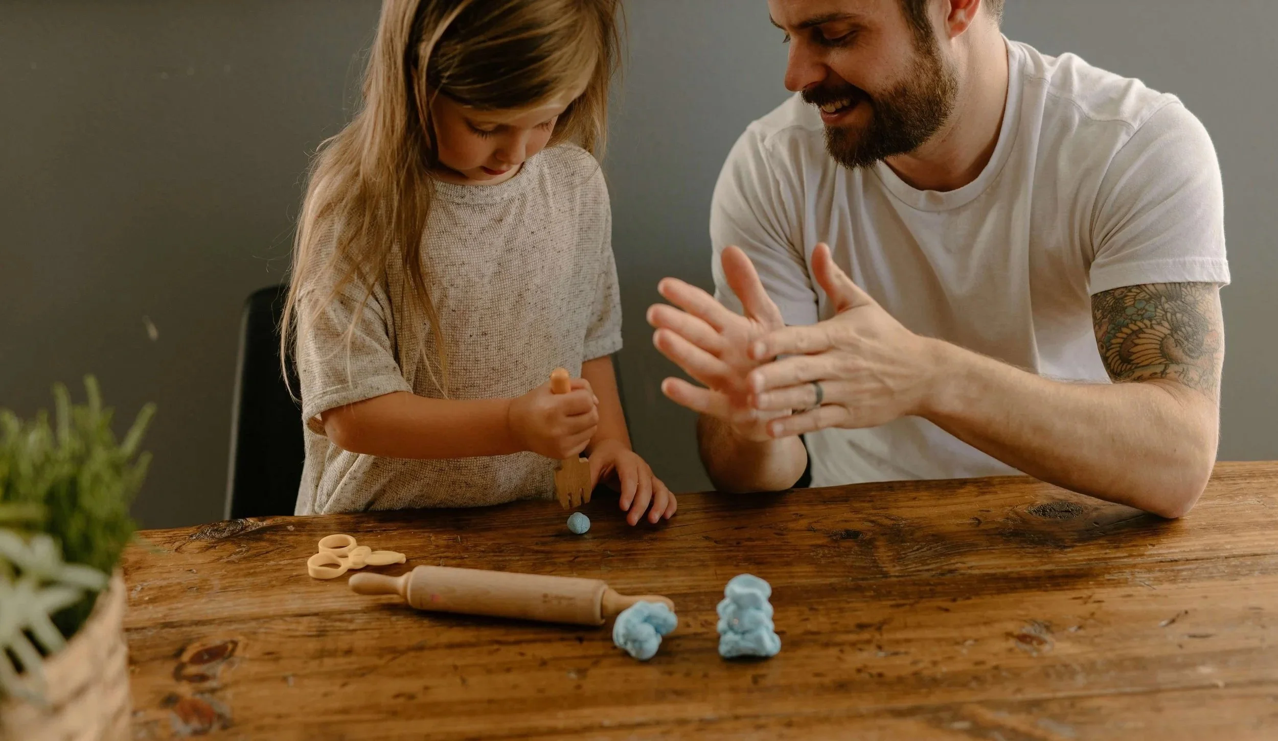 A man and a young girl are playing together at a wooden table with modeling clay and tools. The man has a beard, tattoos on his arm, and is smiling while clapping his hands. The girl has long hair, is wearing a beige shirt, and is focusing on her clay creation.