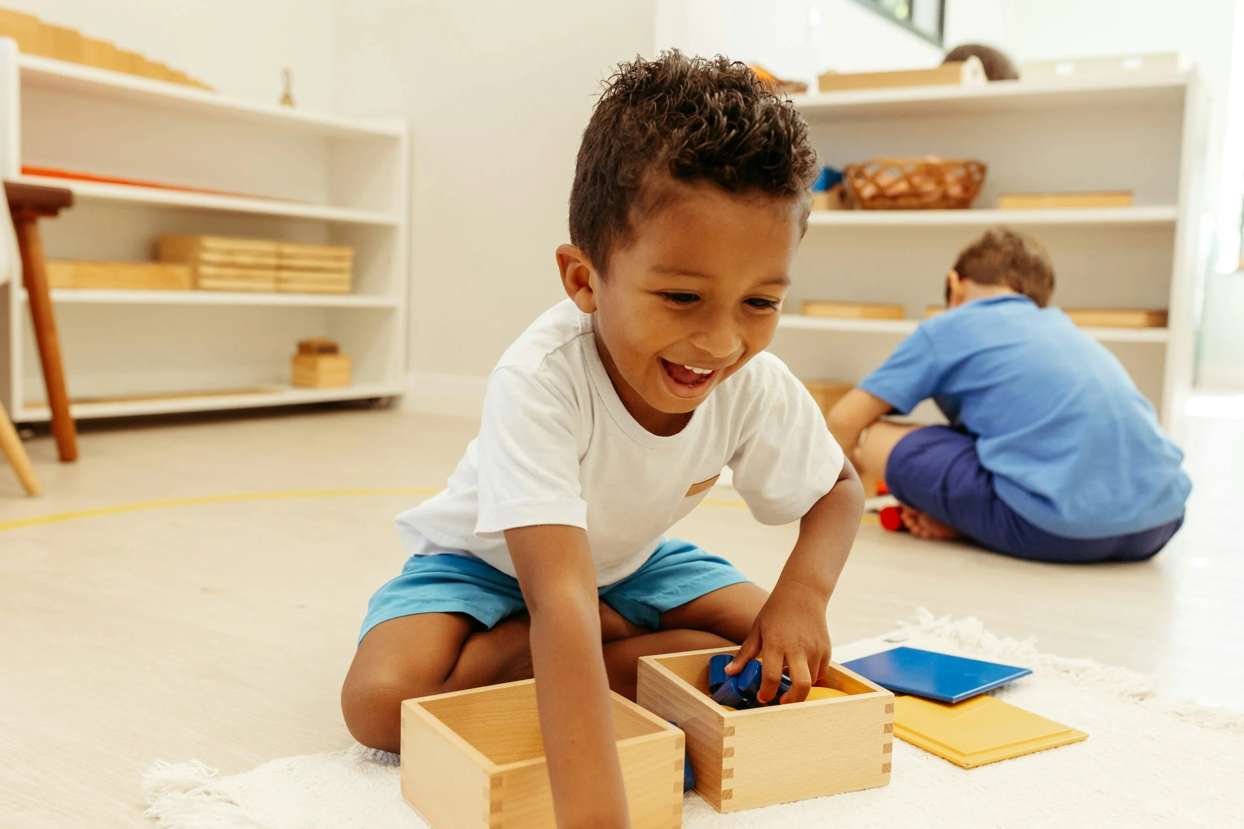 A young boy with curly hair wearing a white t-shirt and blue shorts, smiling and playing with wooden toys on the floor, while another boy wearing a blue shirt and dark shorts is seen in the background.