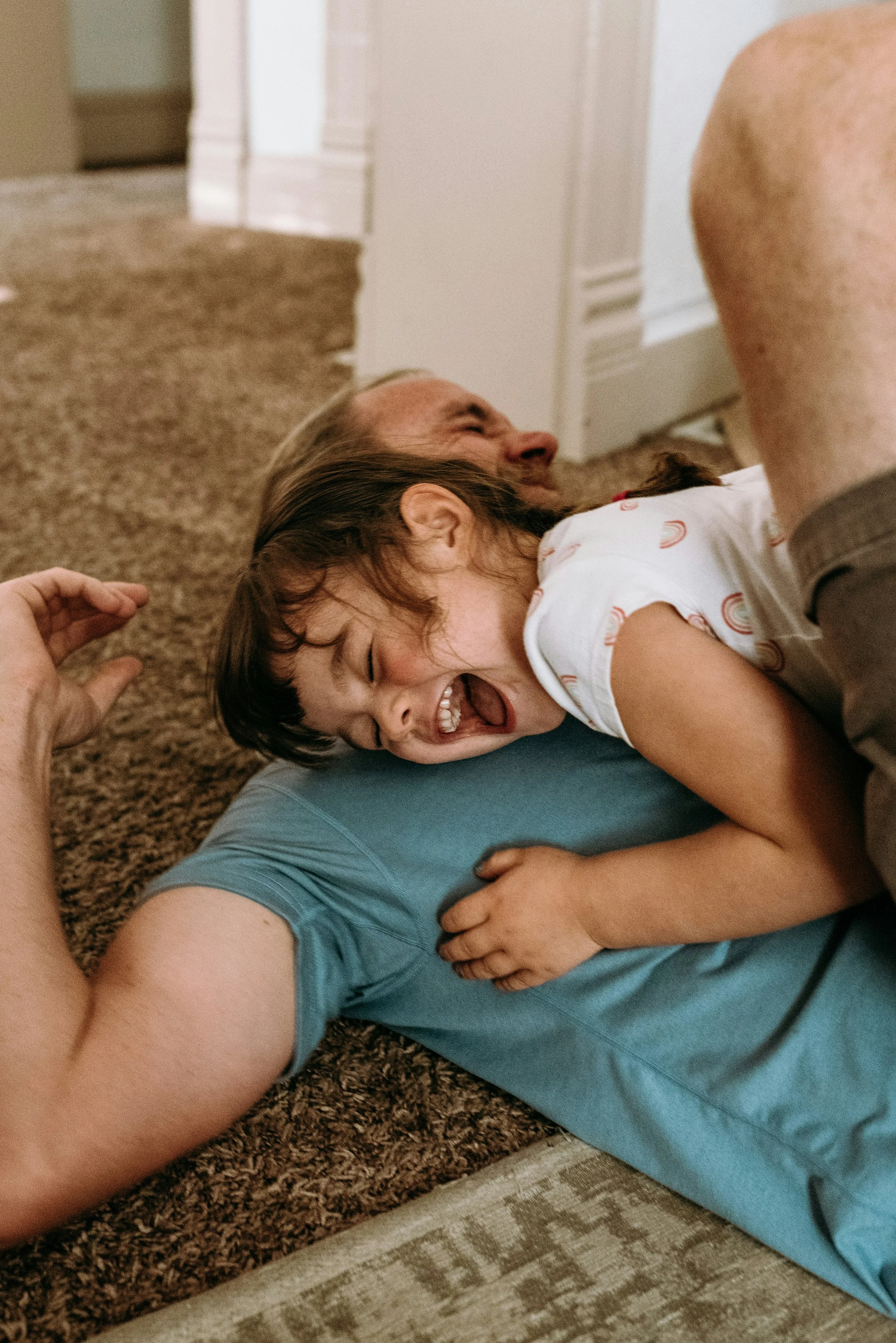 A child and parent lay on the floor. The child is giggling, laying on the parent's chest.