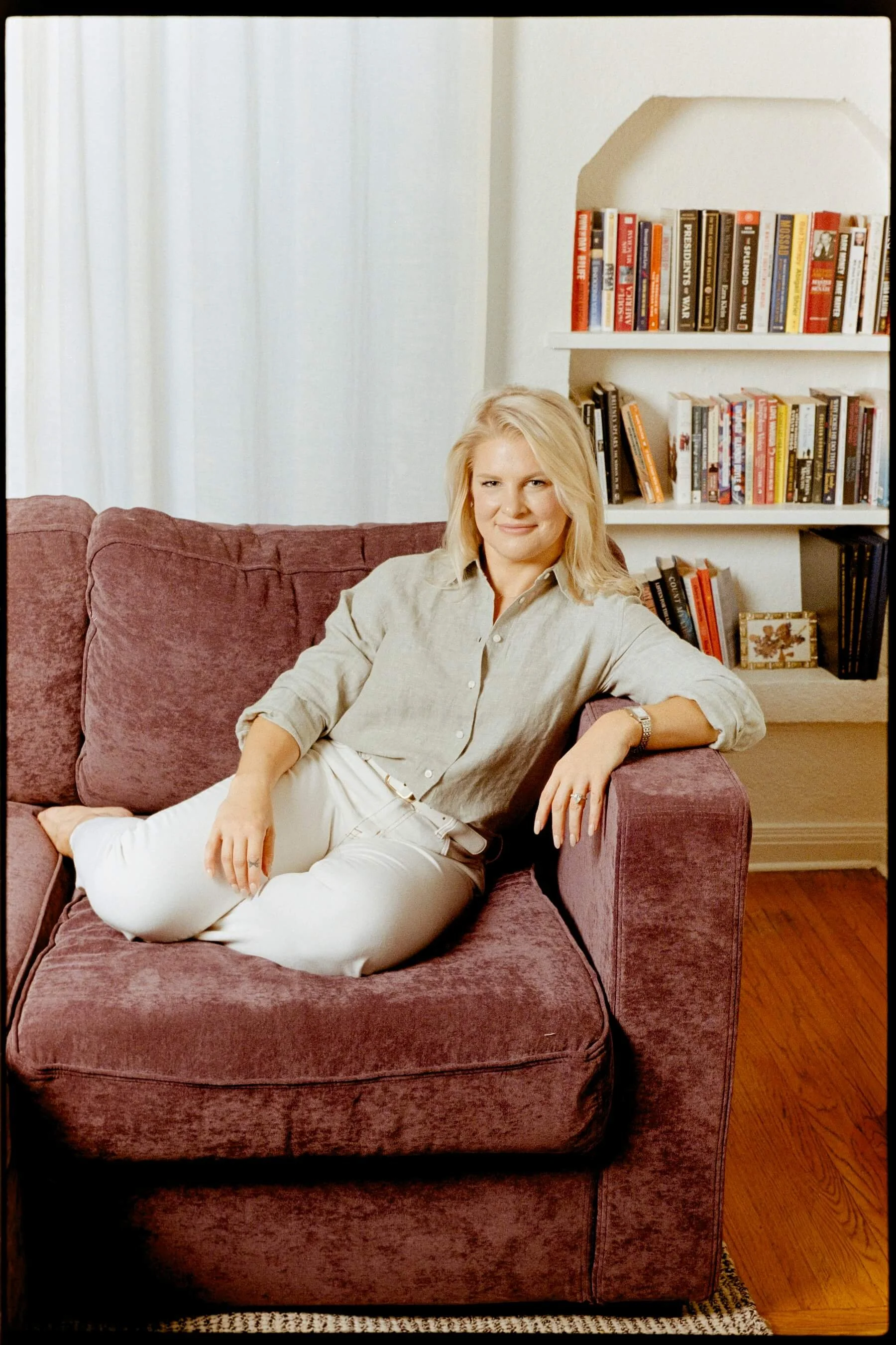 A woman with blonde hair and fair skin sitting on a maroon velvet sofa in a living room with a white bookcase filled with books behind her, wearing a beige shirt and white pants, with her legs crossed and smiling at the camera.