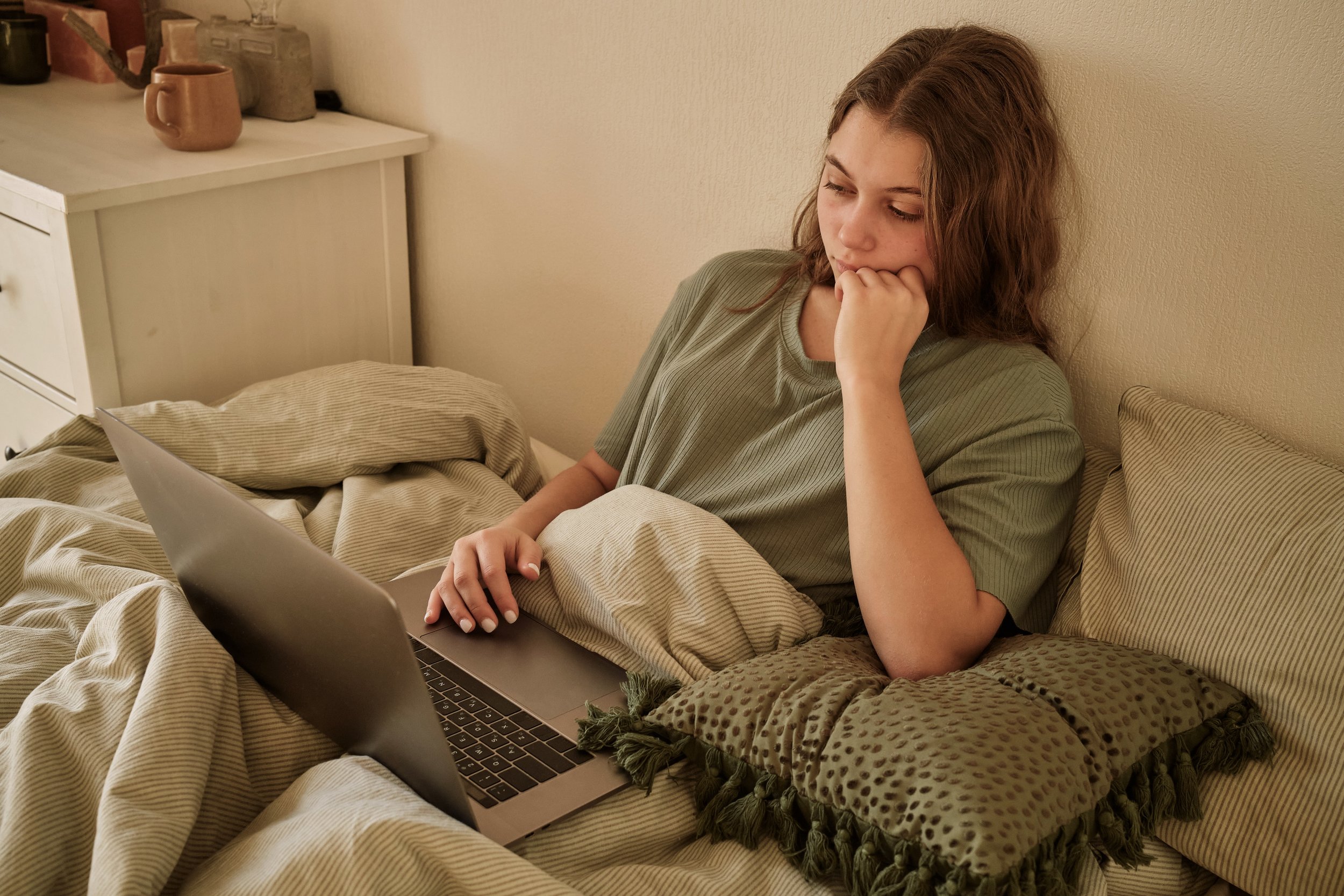 A young woman with curly brown hair sitting on a bed, using a laptop. She is wearing a greenish-gray T-shirt and is resting her chin on her hand, appearing thoughtful.