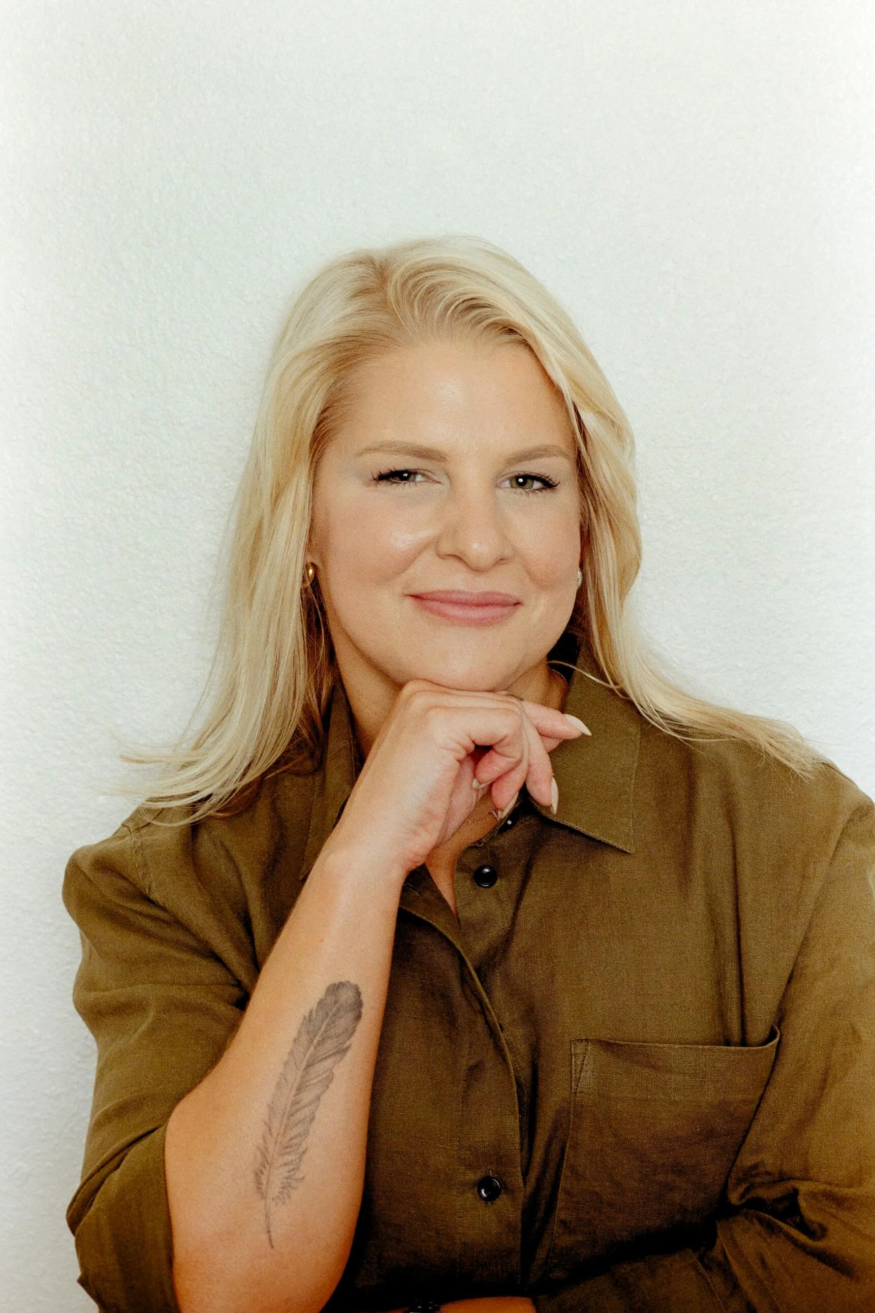 Woman with blonde hair in a brown shirt, resting her chin on her hand, with a tattoo of a feather on her forearm and earrings, standing against a plain white wall.