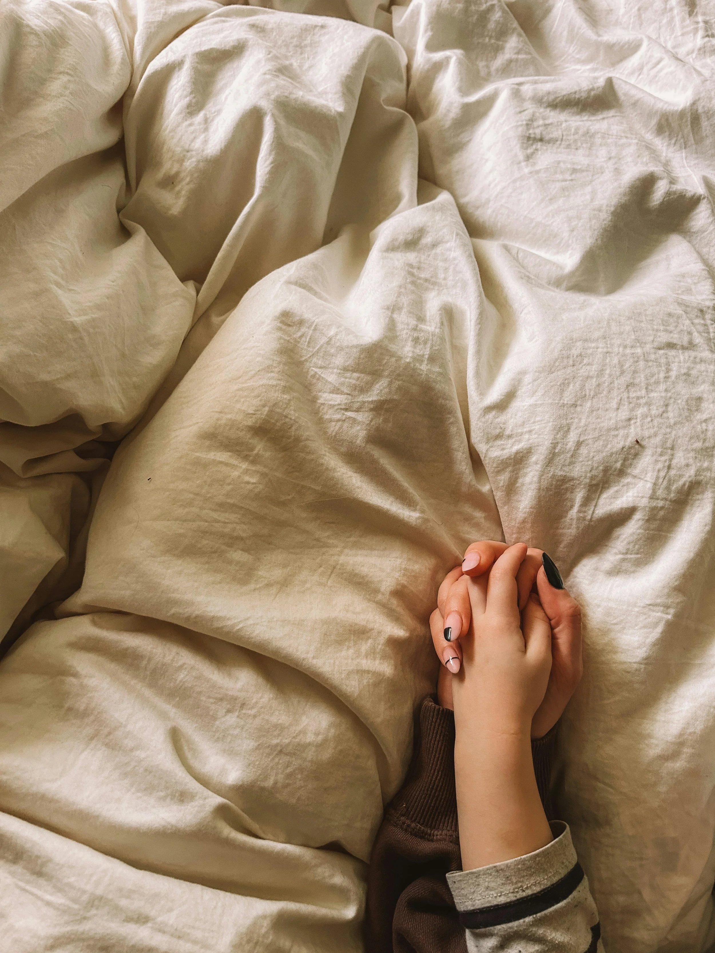 A person with nails painted dark green holding a hand with a black ring, lying on a beige bed with cream-colored sheets.