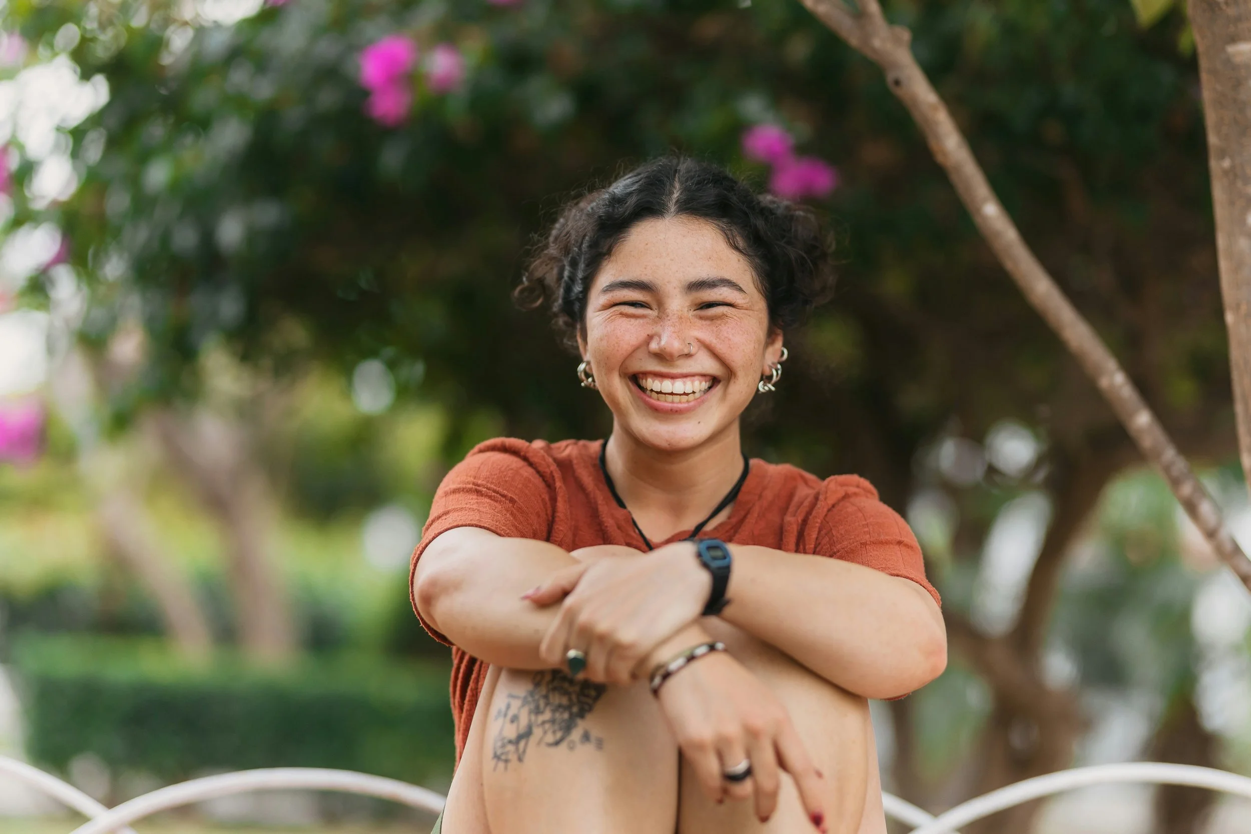 A young woman with short curly dark hair, smiling broadly, sitting outdoors in a park with her arms crossed over her knees. She is wearing a rust-colored shirt, multiple earrings, and a nose ring, with a tattoo on her thigh visible. Blurred greenery and pink flowers are in the background.