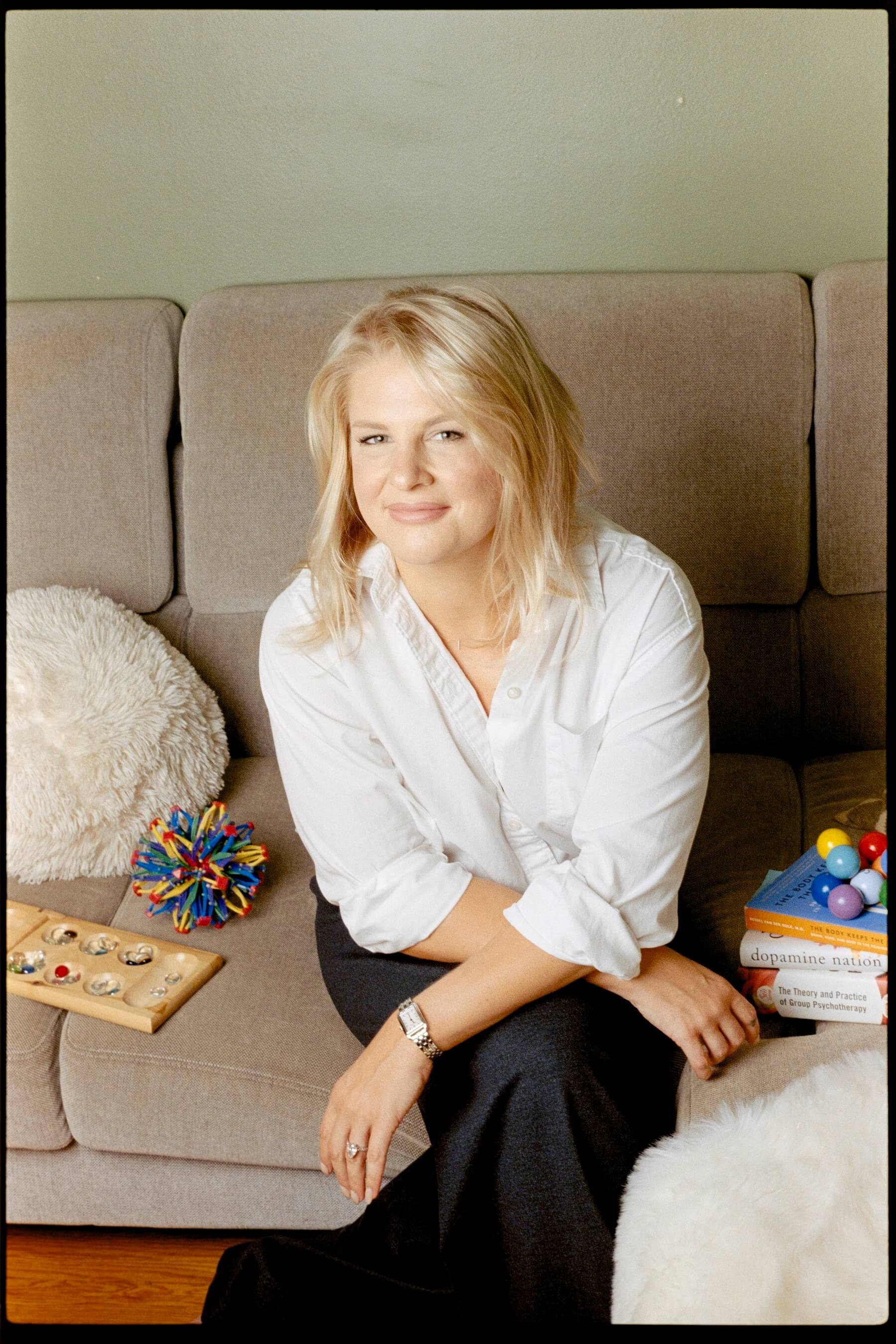 A woman with blonde hair sitting on a beige sofa surrounded by books and toys, including a colorful spiky ball, a wooden puzzle, and a box of multicolored balls. She is wearing a white shirt and black pants.
