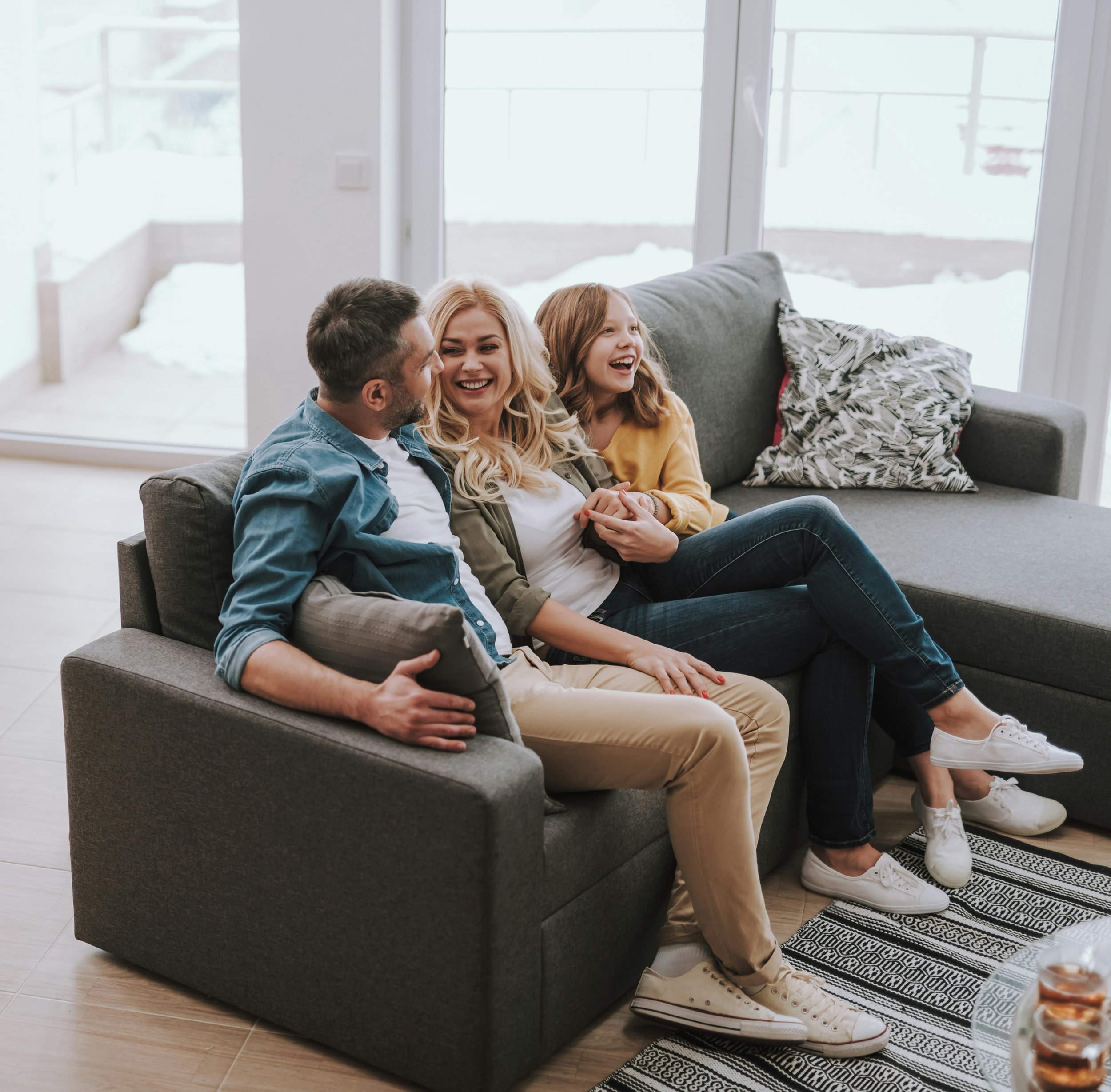 A family of four sitting on a gray couch near large windows inside a modern home. The family appears to be enjoying each other's company, with smiles and laughter. The room has a casual, cozy atmosphere with a patterned rug and a decorative pillow.