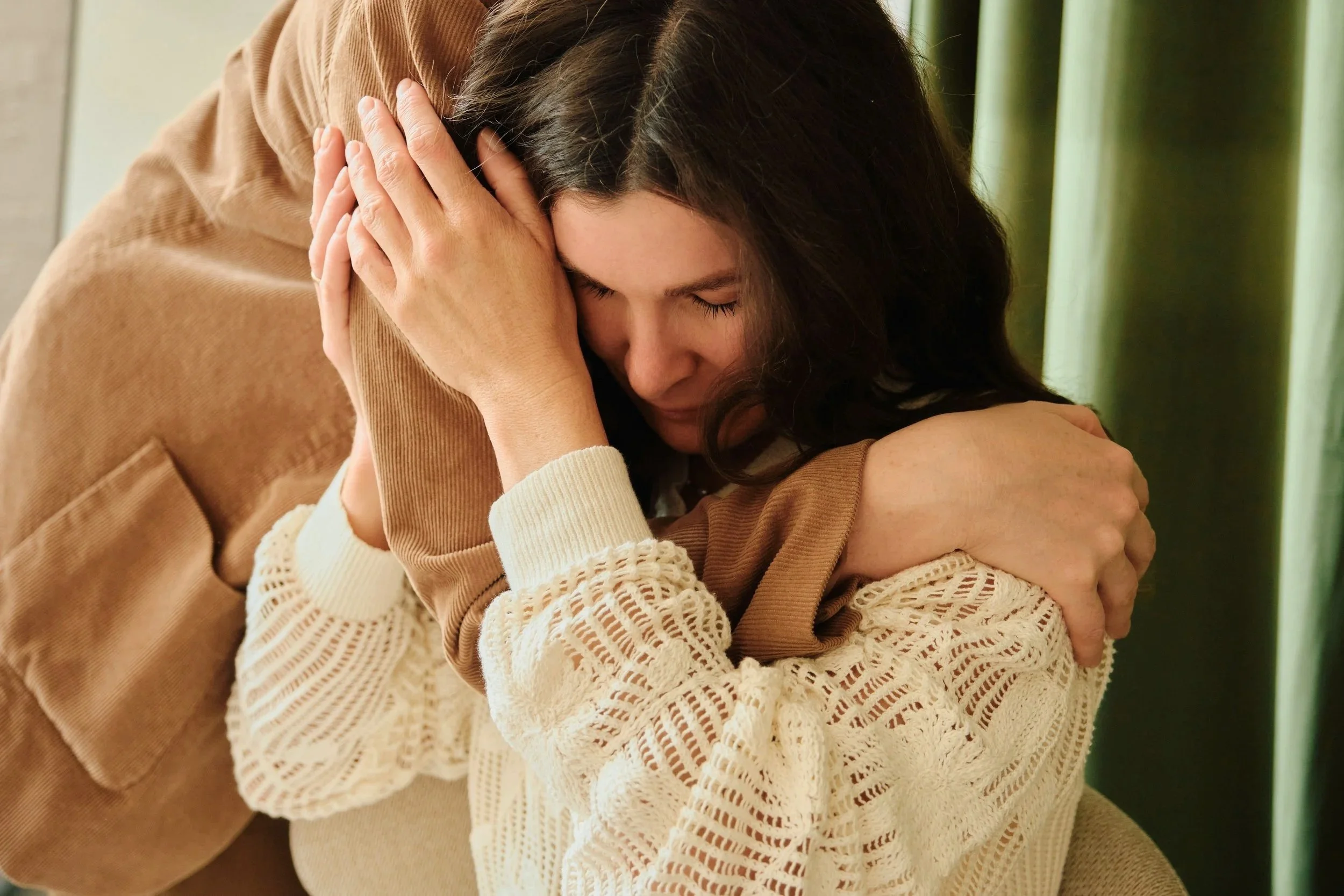 A woman in a cream-colored knitted sweater hugging a person in a brown jacket with knots in the back. The woman's face shows emotion, with her eyes closed and her head resting on her arms.