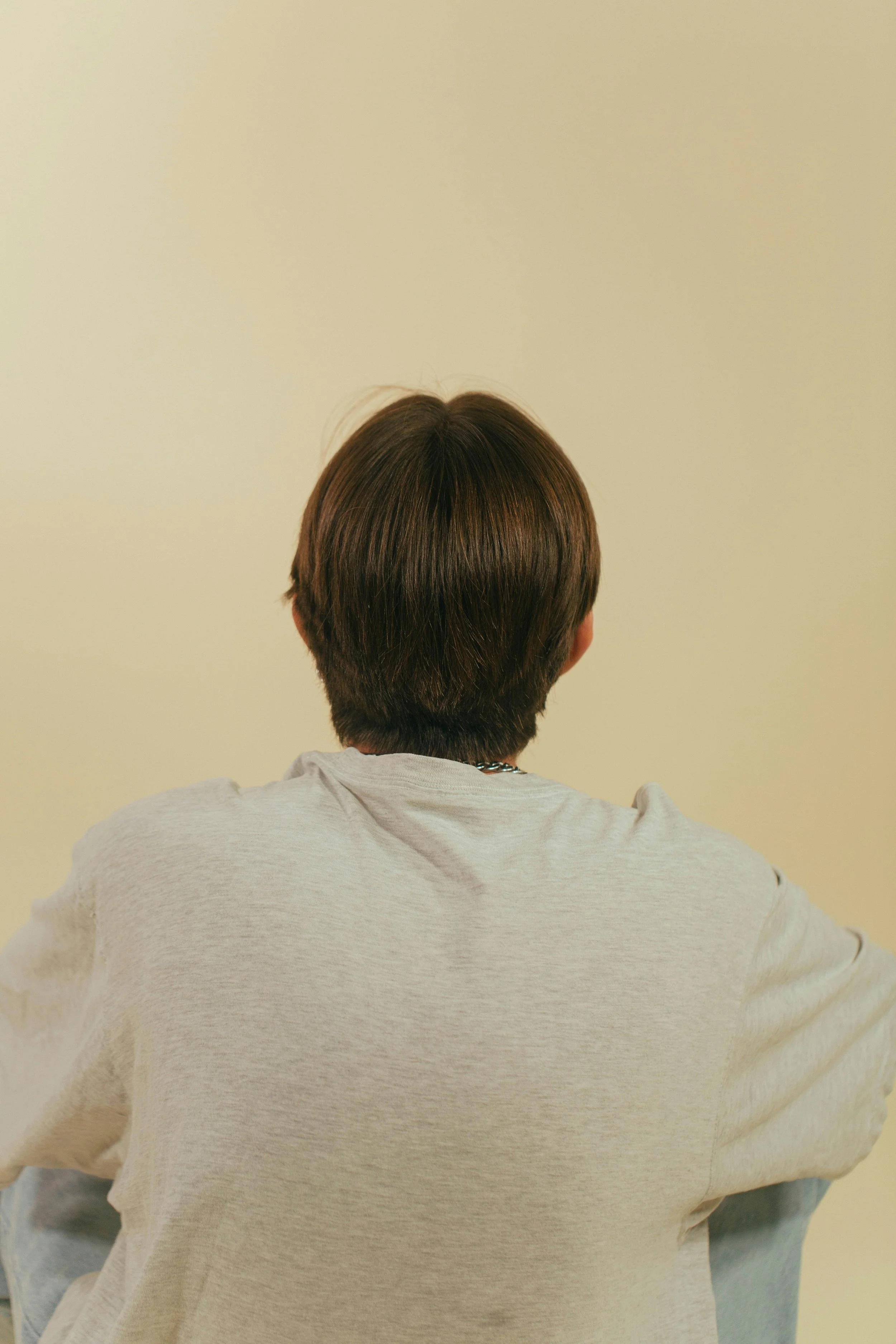 Person with short brown hair wearing a light-colored shirt, viewed from behind, sitting on a yellow wall.