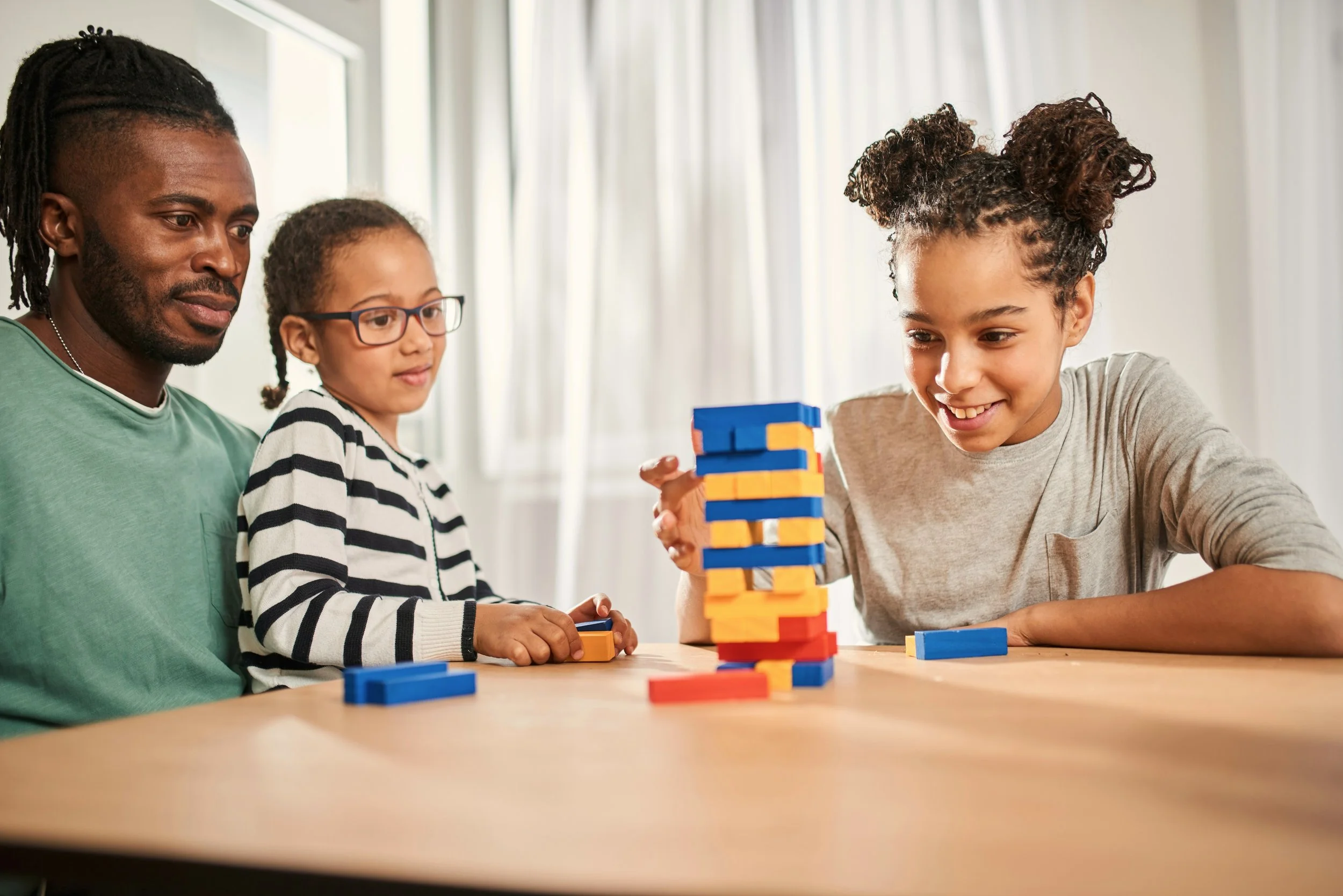 Three people, two children and an adult, playing a game with colorful blocks at a wooden table in a bright room.