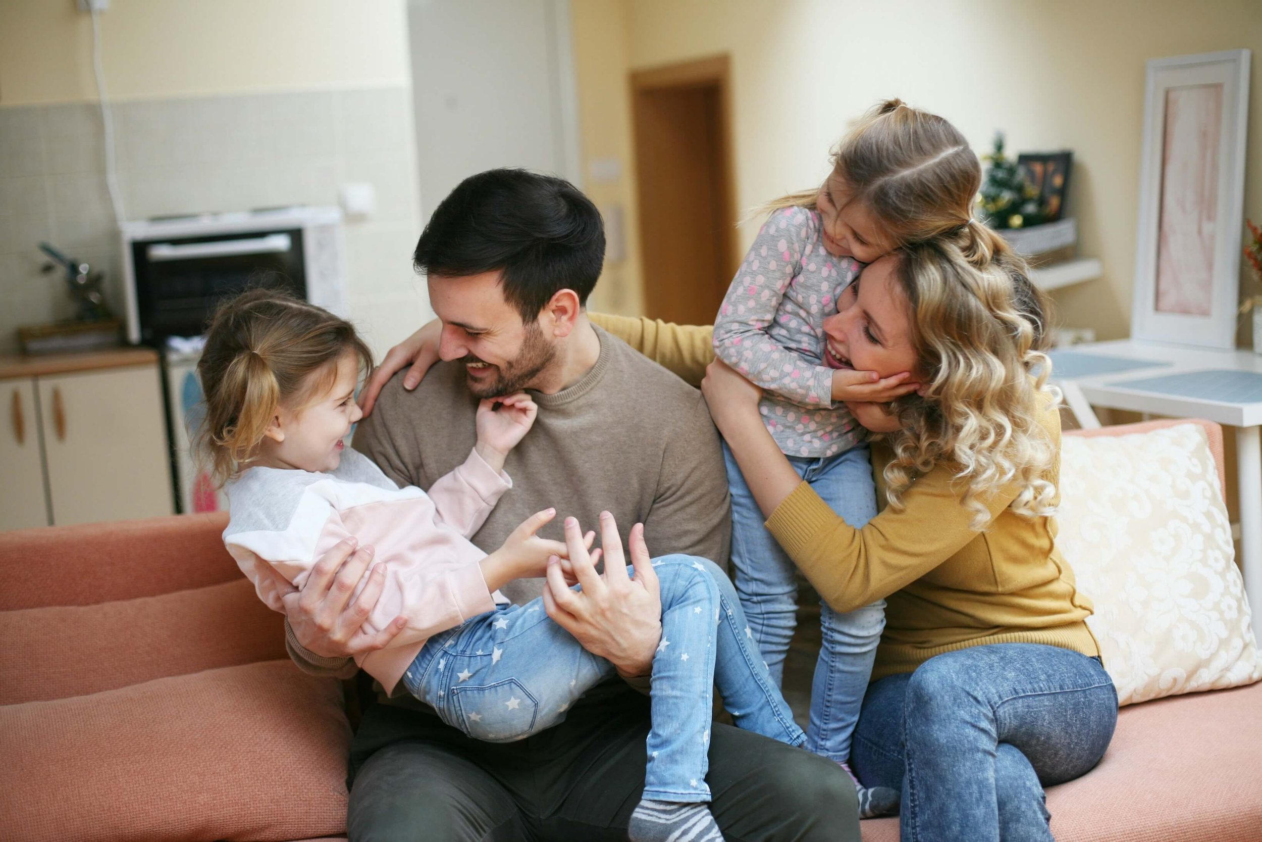 A family of five, including two young girls, is sitting on a sofa in a cozy living room, laughing and playing together.