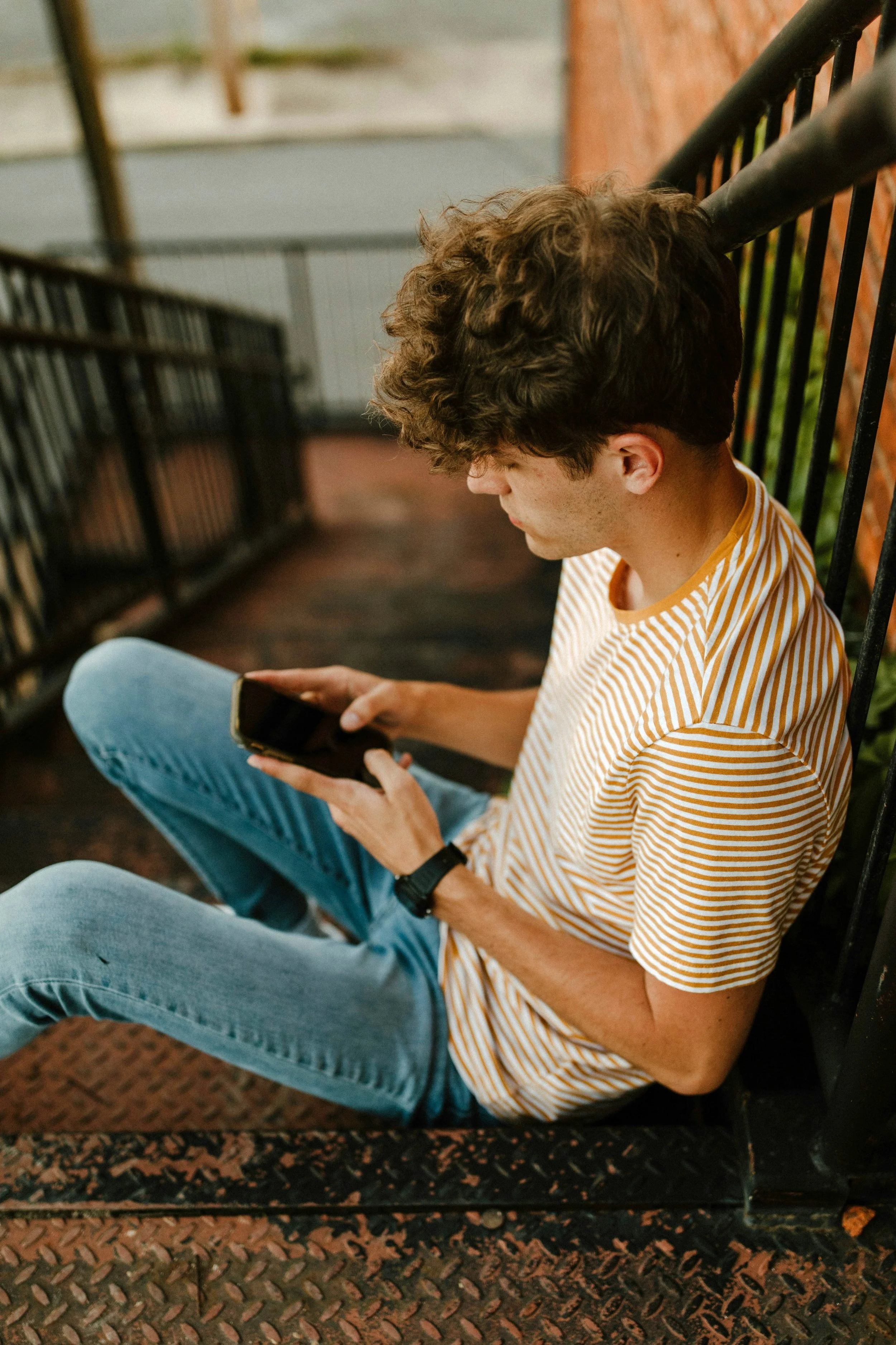 A young man with curly brown hair sitting on a metal stairway, looking at a smartphone, wearing a striped yellow and white T-shirt and blue jeans.