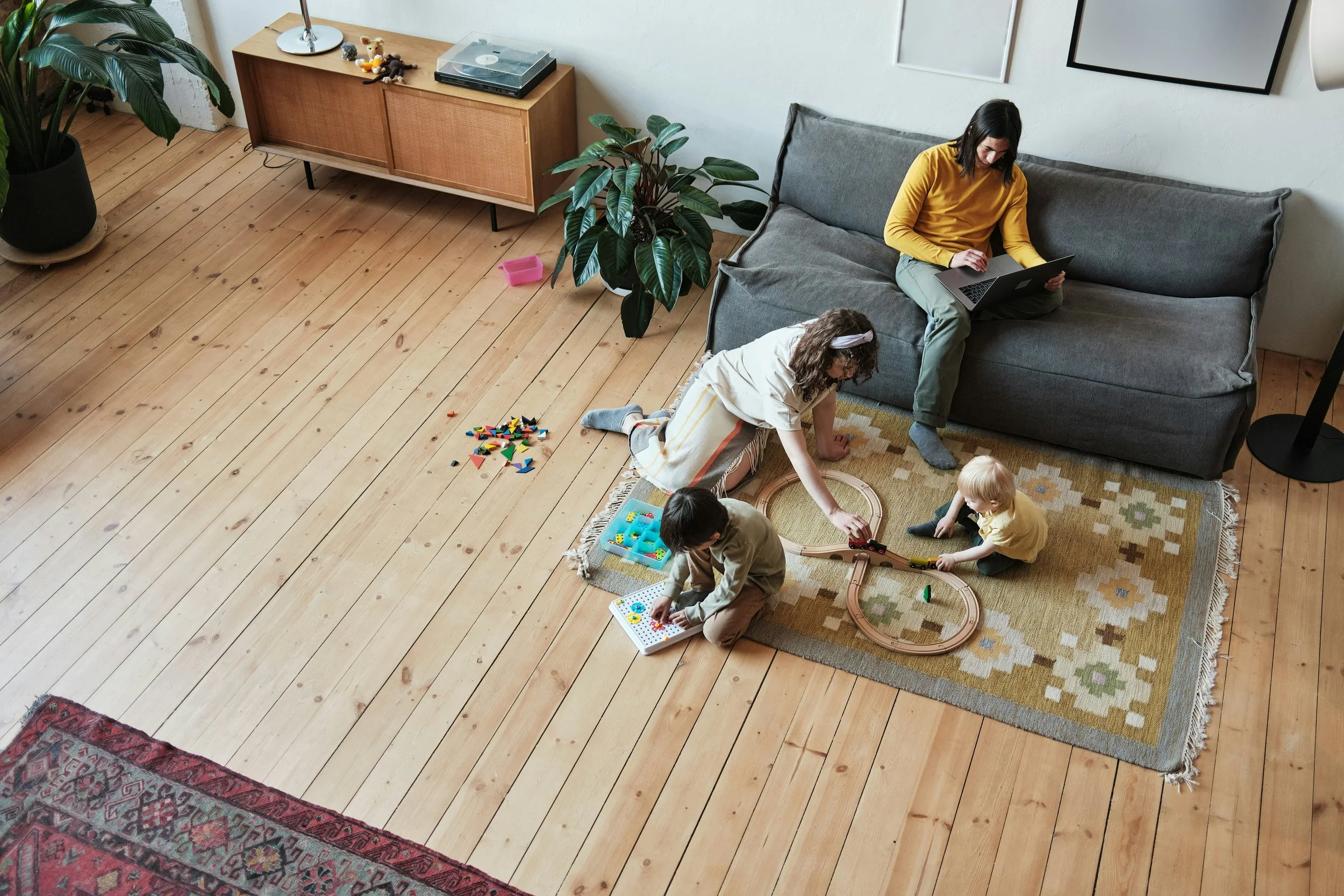 A woman sitting on a gray couch working on a laptop, with two children playing on a rug on the wooden floor nearby. One child is kneeling and the other is sitting, both engaged with toys and a train set.