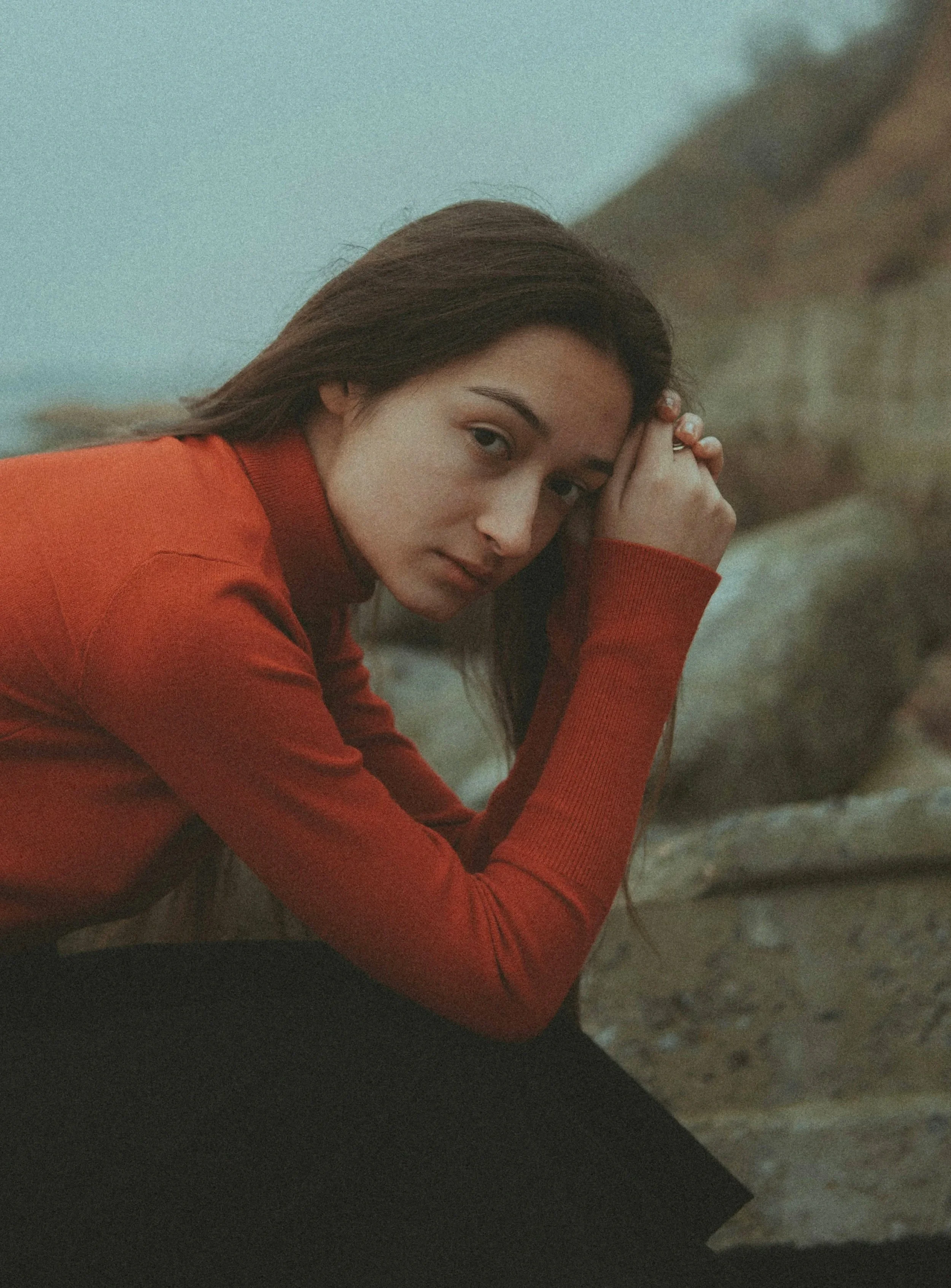 A woman with long brown hair wearing a red sweater, resting her head on her hand, looking at the camera with a calm expression on a beach with rocky cliffs in the background.