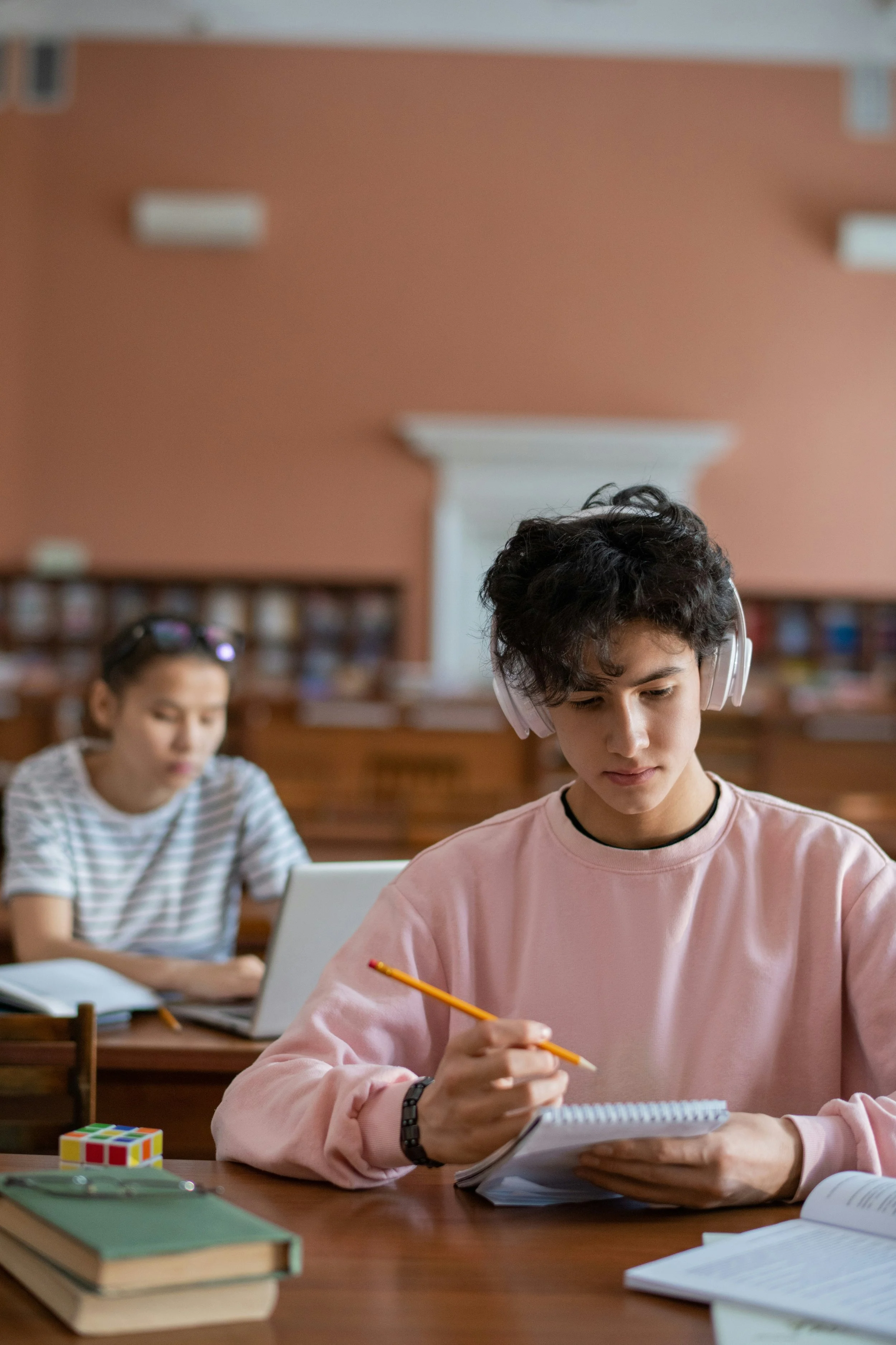 Three students studying in a classroom, one wearing headphones and writing in a notebook, with books and a Rubik's cube on the desk.