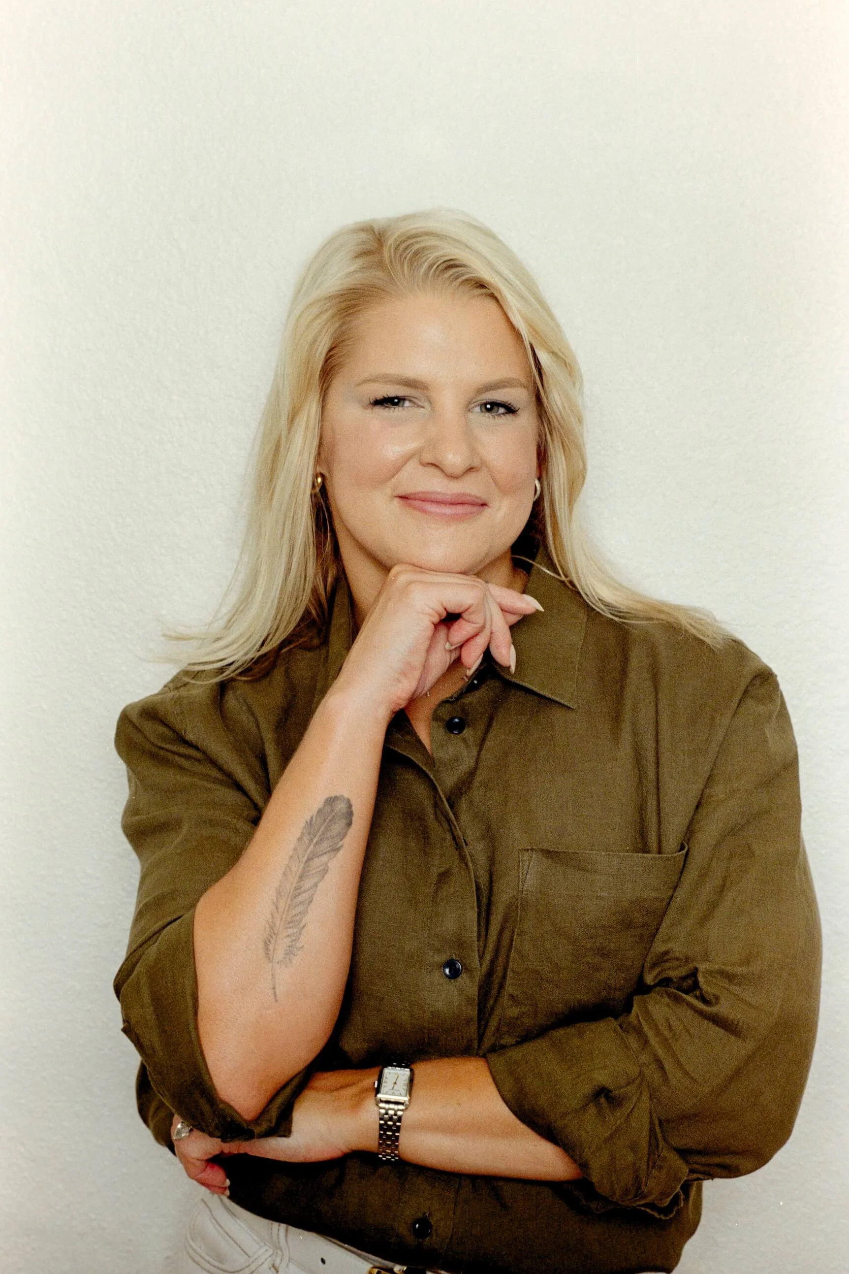 A woman with long blonde hair, wearing a brown shirt and a watch, poses with her hand on her chin and a content smile on her face. She has a feather tattoo on her forearm and is standing against a plain white wall.