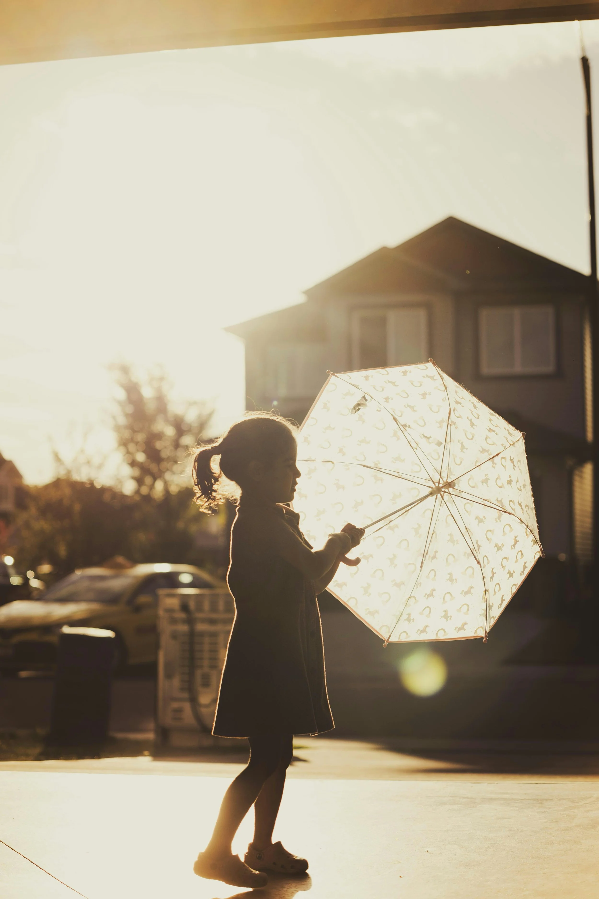 A young girl holding an umbrella with a pattern, walking on a sidewalk during sunset, with the silhouette of a house and trees in the background.