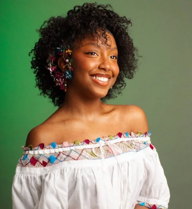A smiling woman with curly black hair decorated with colorful earrings and a white off-the-shoulder top with pom-pom trim, standing against a green background.