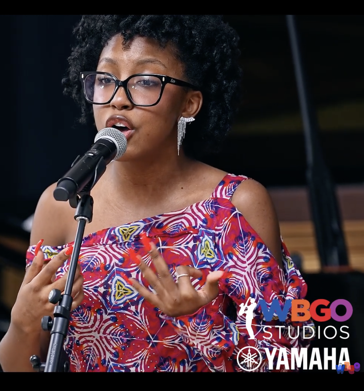 A woman with curly black hair, wearing glasses and a vibrant patterned off-the-shoulder dress, singing into a microphone with her hands gesturing expressively. Logo of #WBG Studios and Yamaha in the bottom right corner.