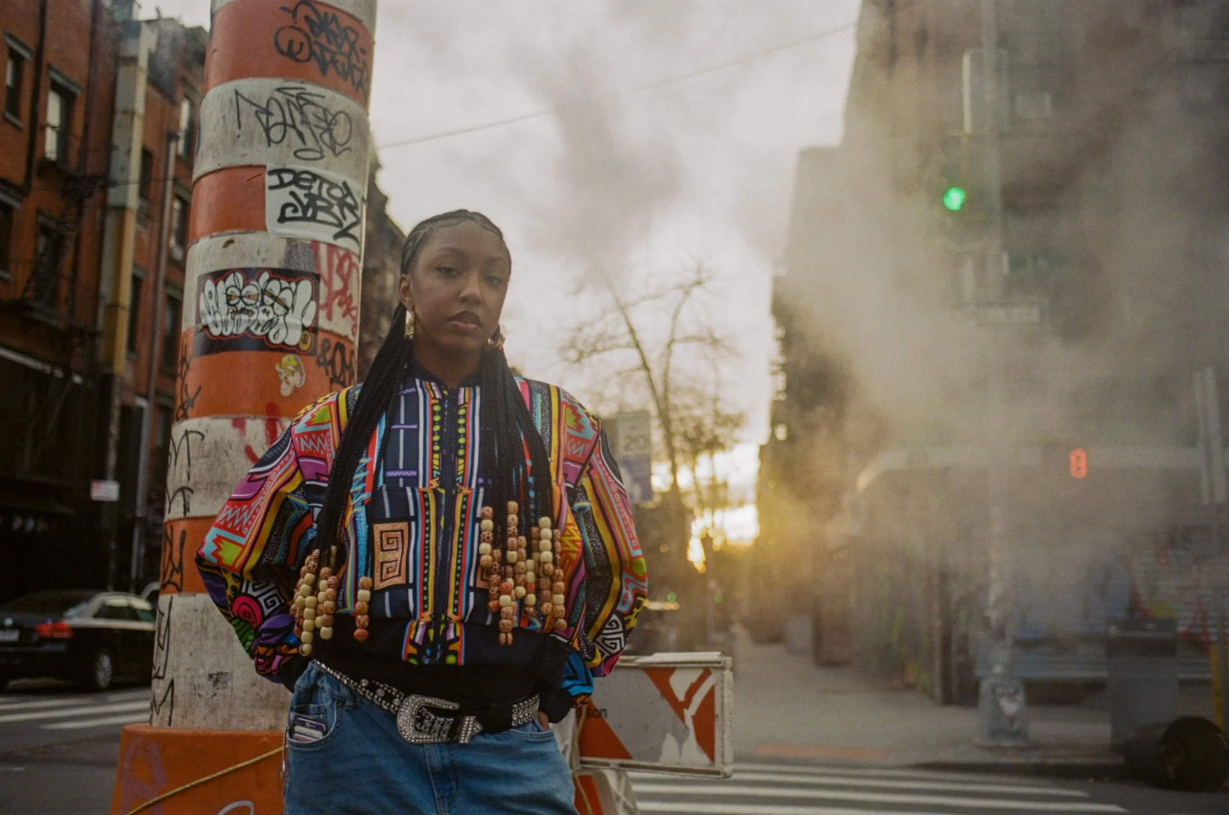 A woman standing on a city sidewalk at dusk, wearing a colorful patterned jacket and multiple beaded necklaces, with her hands in her pockets and a serious expression. Behind her, there is a pole covered in graffiti and a construction barrier. The background shows urban buildings, leafless trees, and some streetlights, with a smoky or foggy atmosphere.