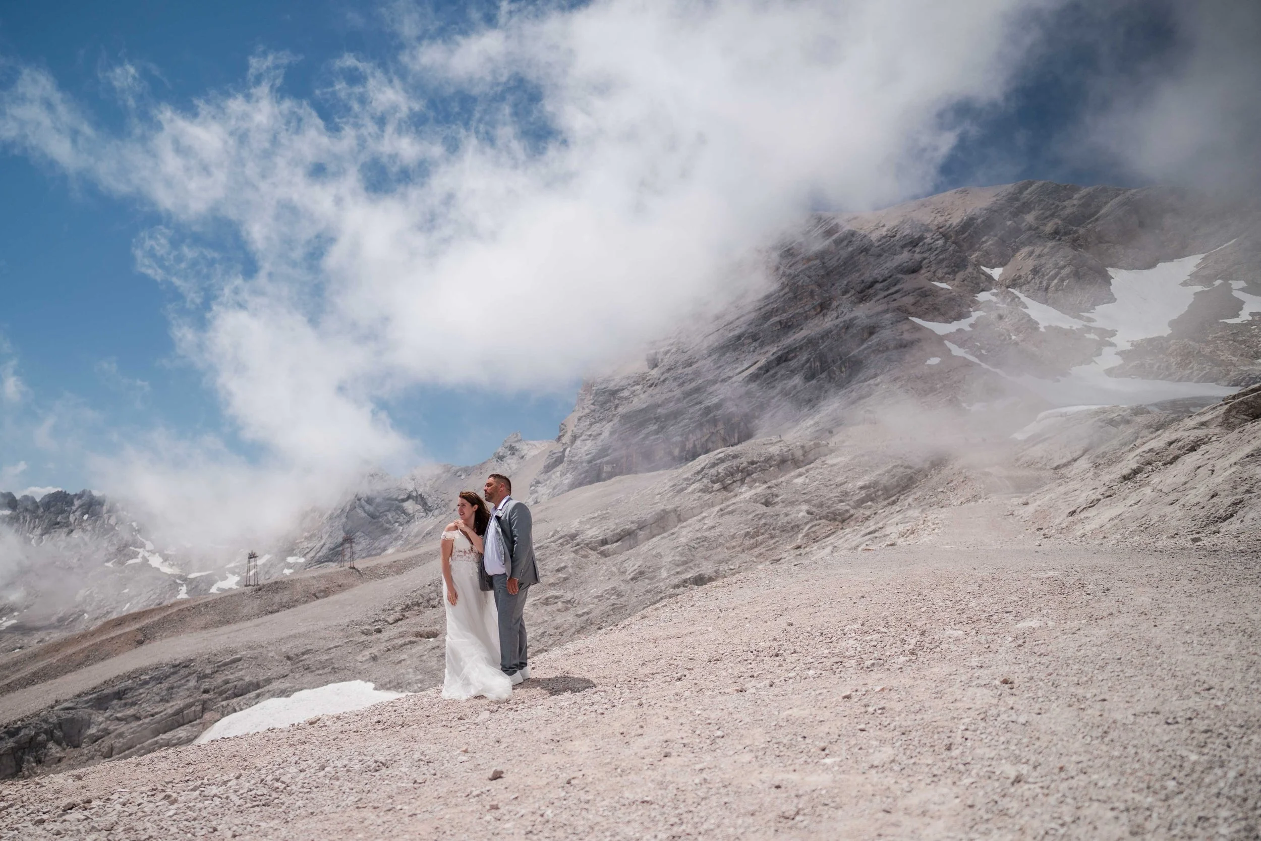 Ein Brautpaar in Hochzeitskleidung steht in einer felsigen, bergigen Landschaft mit Himmel und Wolken im Hintergrund.