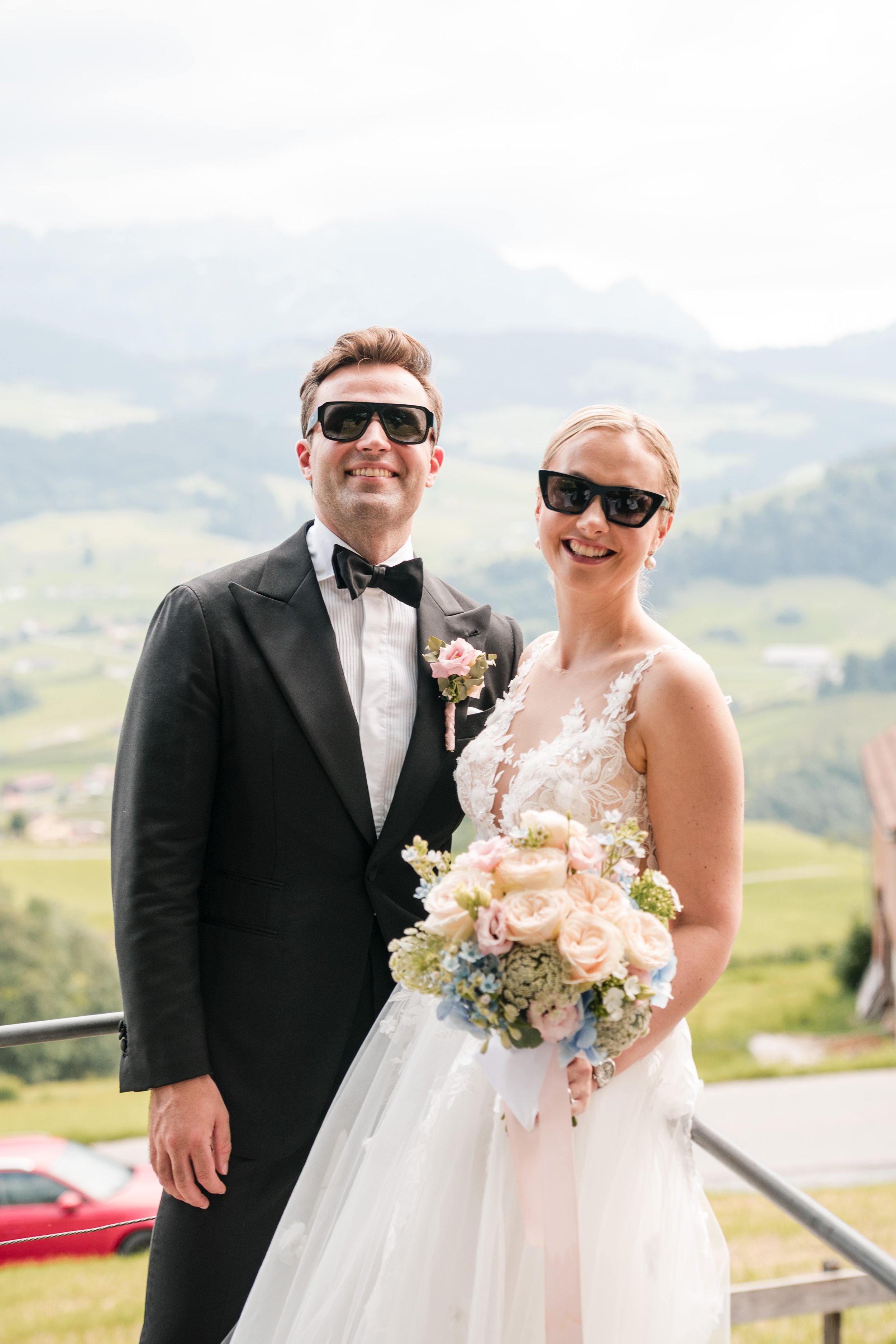 Braut und Bräutigam in Straßenkleidung mit Sonnenbrillen, vor grünem Hügel- und Bergpanorama, bei einer Hochzeit im Freien.