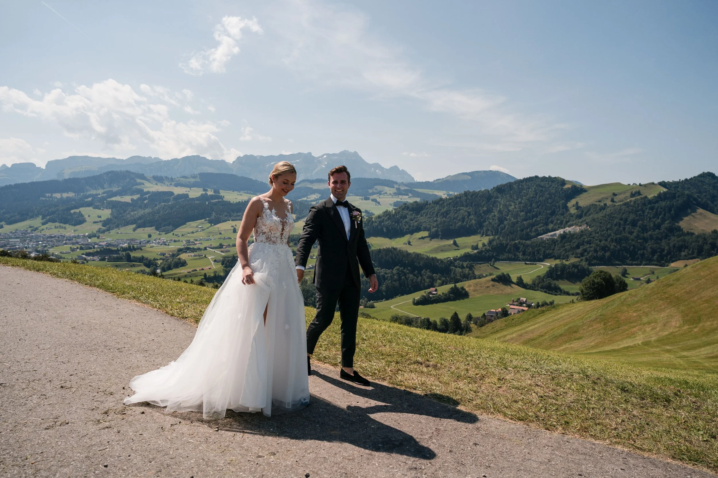 Ein Brautpaar auf einer Wiese mit Blick auf grüne Hügel und Berge unter einem sonnigen Himmel.