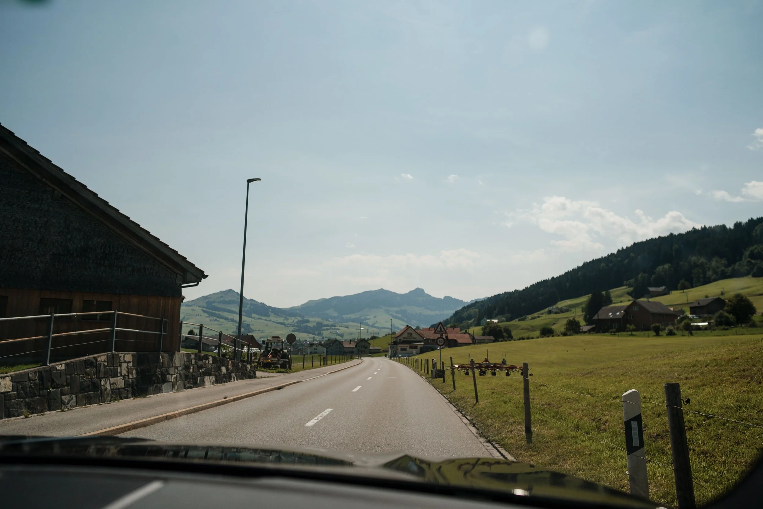 Eine ländliche Straße, umgeben von grünen Wiesen und Hügeln, mit Bergen im Hintergrund, unter einem überwölbten Himmel.
