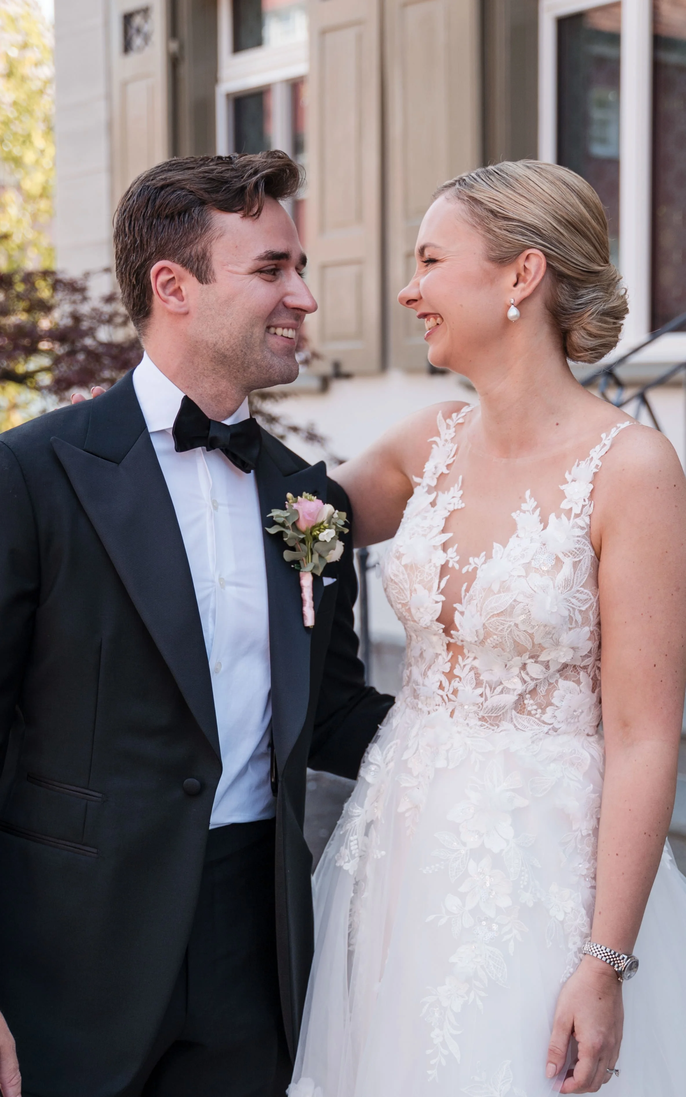 Ein Brautpaar lächelt sich an, sie trägt ein weißes Hochzeitskleid mit Spitzenmuster, er trägt einen schwarzen Smoking mit Fliege, draußen vor einem Haus.