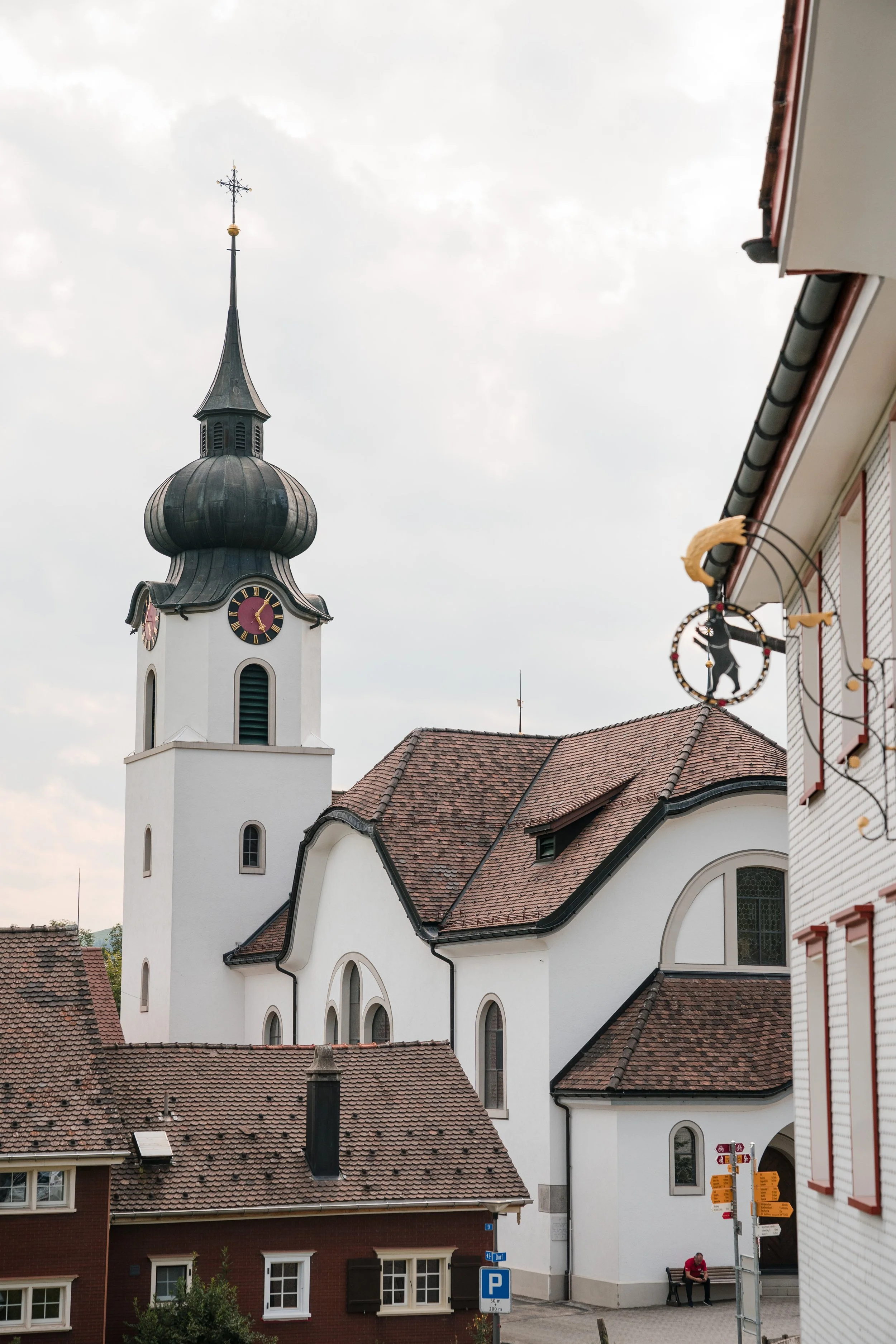 Eine weiße Kirche mit dunklem, geschwungenem Dach und einem Uhrturm mit Glocken, vor einem bewölkten Himmel. Im Vordergrund sind Dächer und eine Person, die auf einer Bank sitzt, sichtbar.