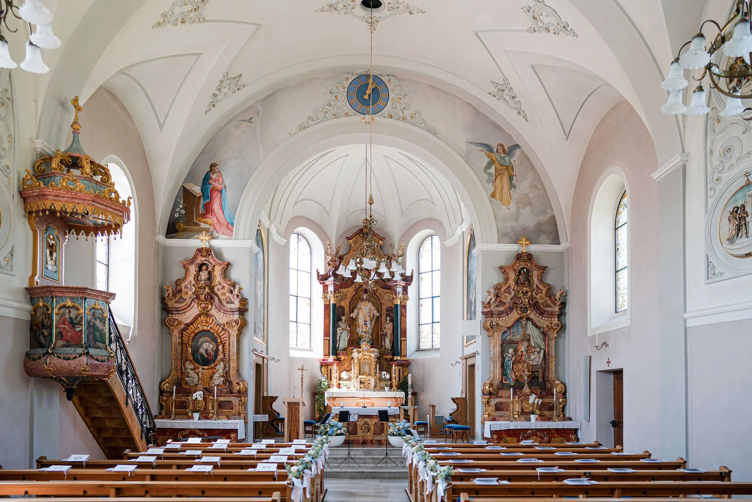 Innenansicht einer barocken Kirche mit Altar, alten Holzbänken, weißen Wänden, bunten Wandmalereien, Horntüren und hohen Fenstern, das Licht fällt durch die Fenster auf den Boden.
