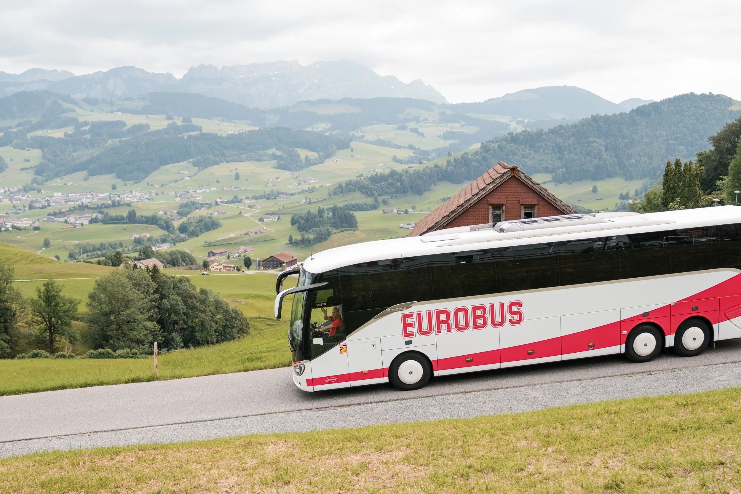 Ein weißer Bus mit rotem Schriftzug 'EUROBUS' fährt auf einer Straße durch eine grüne, hügelige Landschaft mit Häusern und Bergen im Hintergrund.