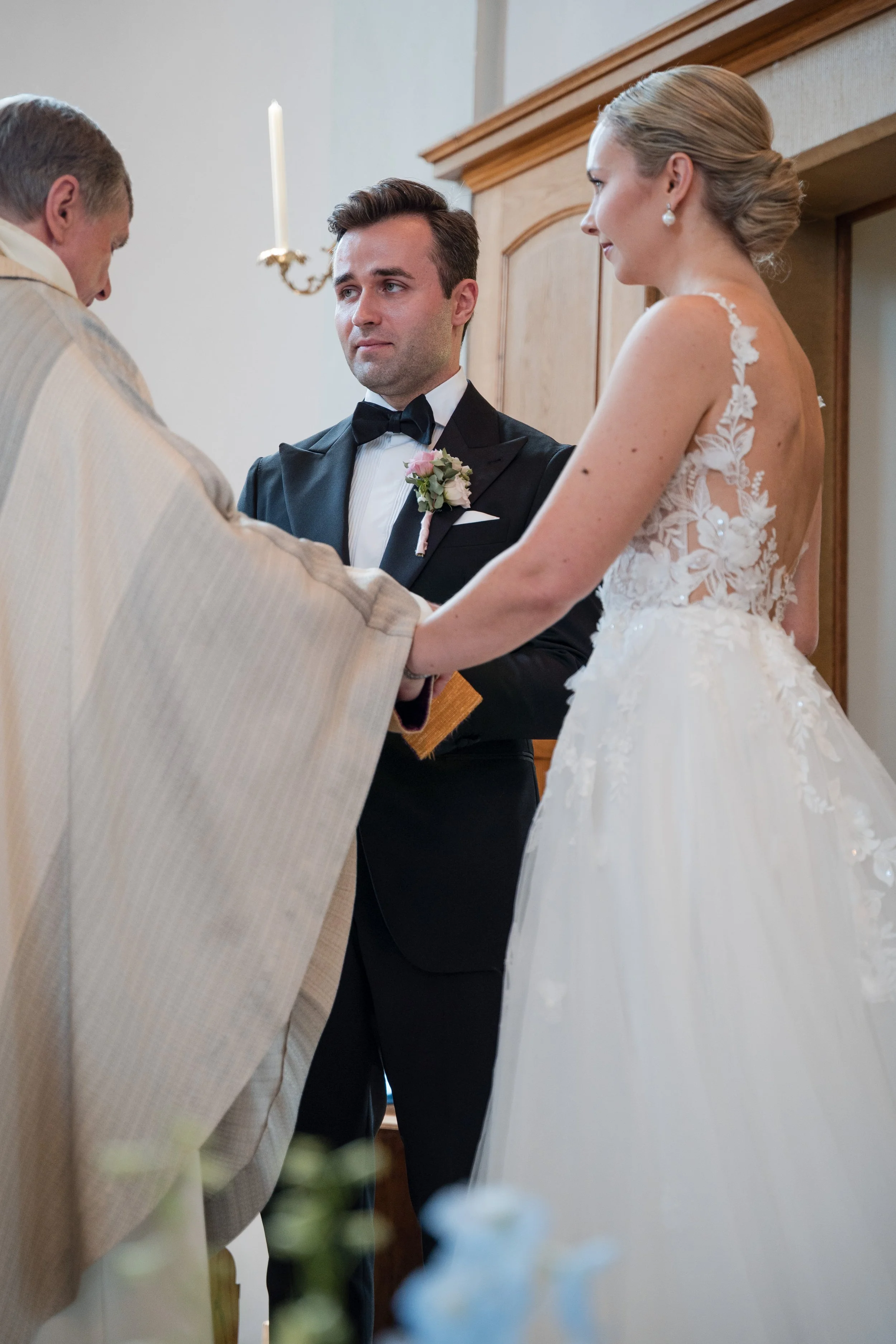 Ein Brautpaar bei der Trauung, der Bräutigam trägt einen schwarzen Anzug mit Fliege, die Braut trägt ein weißes Hochzeitskleid mit floralen Applikationen, sie halten sich an den Händen, der Priester vollzieht die Zeremonie in einer Kirche.