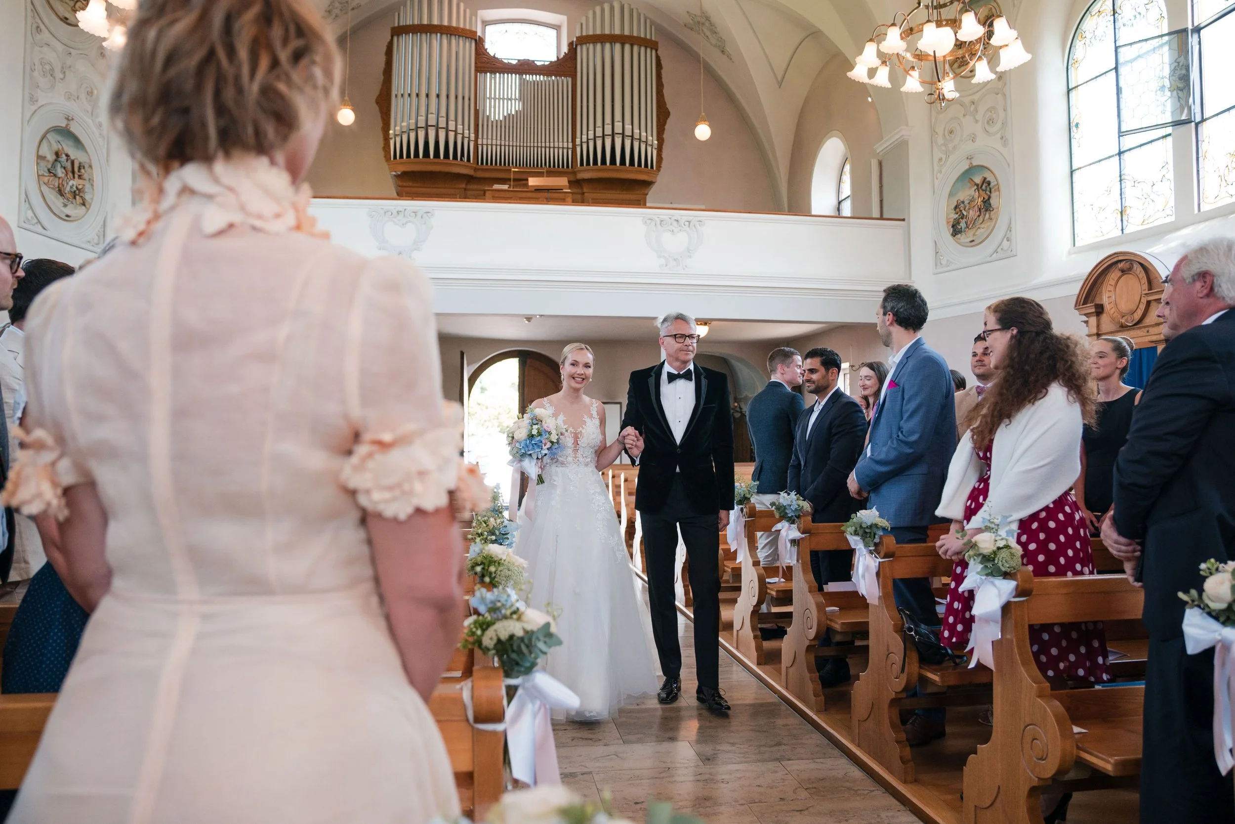 Braut und Vater laufen in einem festlich geschmückten Kirchenraum nach vorne, während Gäste in den Sitzbänken ihnen zuschauen.