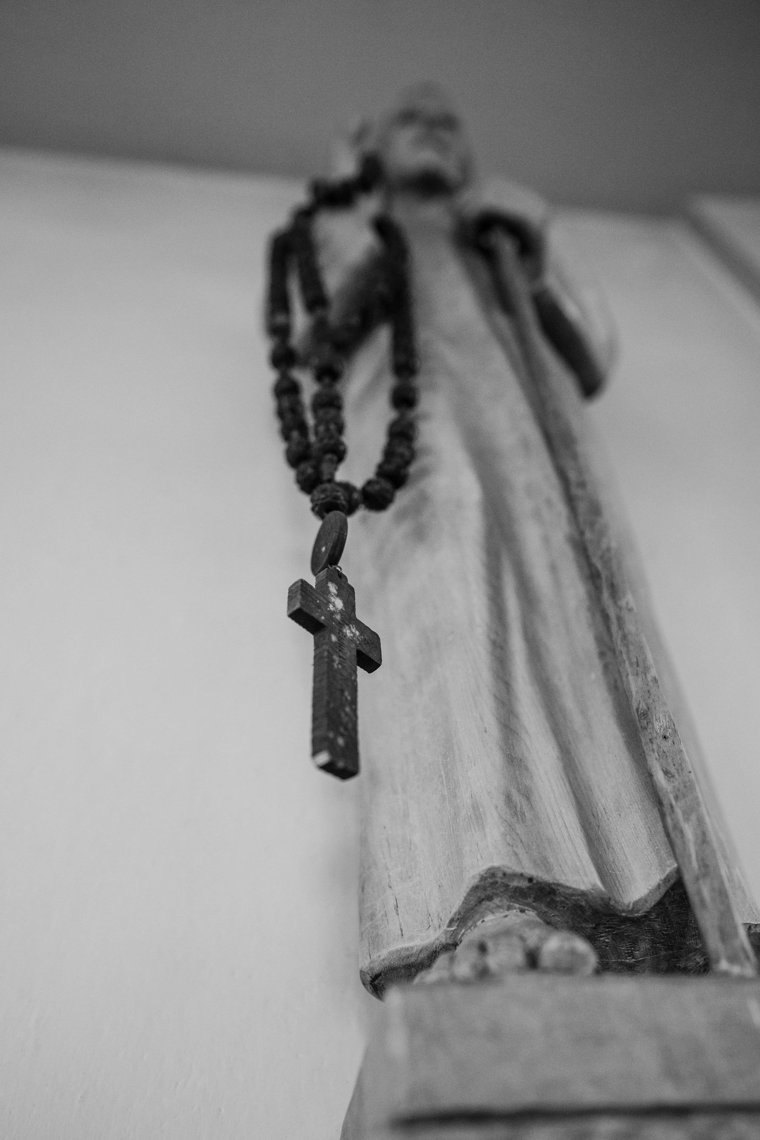 Schwarz-Weiß-Foto einer religiösen Statue, die eine Figur mit einem Kreuz in der Hand darstellt, mit einem Gebetskranz. Die Statue ist auf einem Sockel aufgestellt.