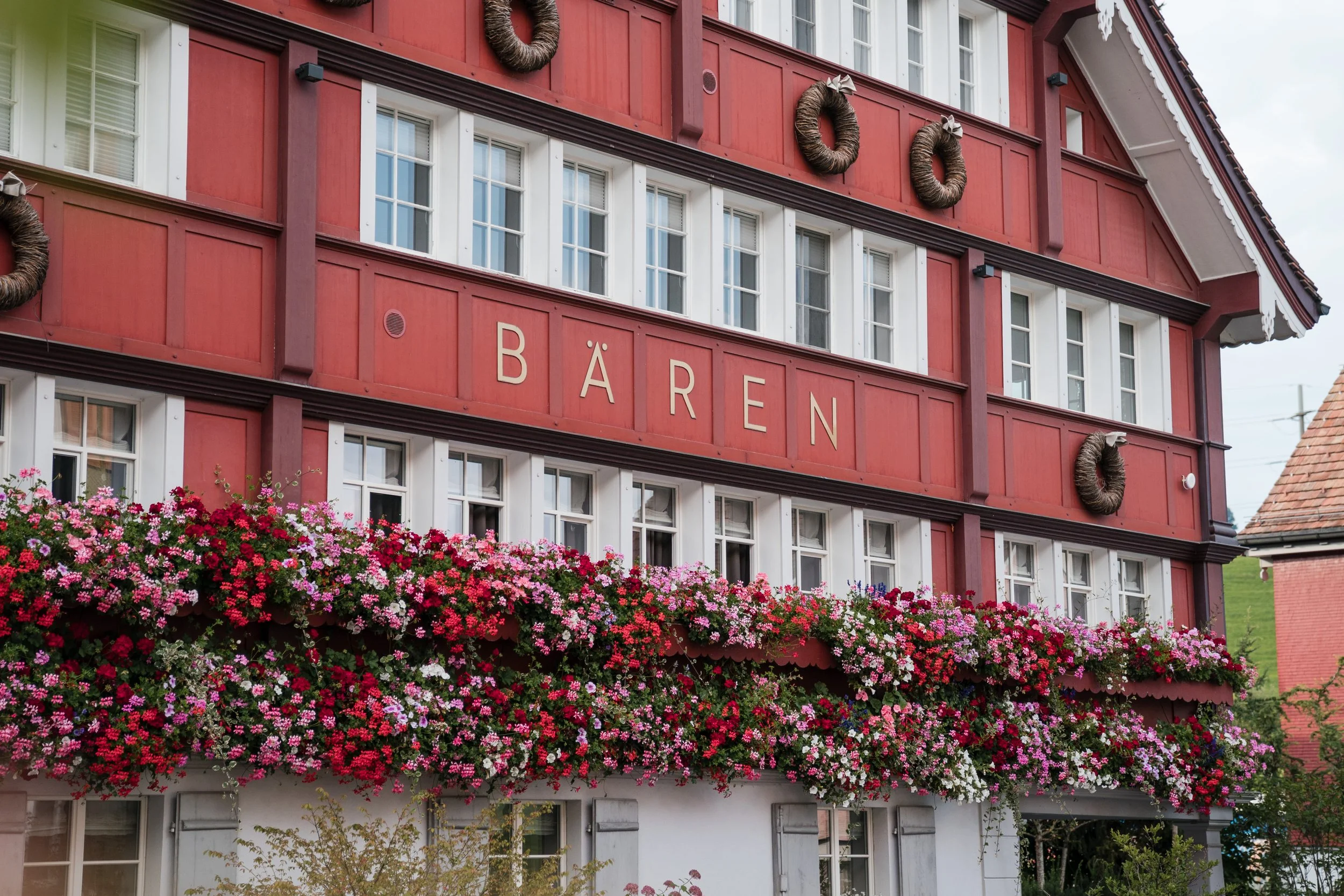 Ein mehrstöckiges, rotes Gebäude mit weißen Fensterrahmen und einem Blumenkasten voller bunter Blumen an der Fassade. Das Wort "Bären" ist auf der Fassade zu lesen, und es sind Dekorationen in Form von Kränzen an der Hauswand angebracht.