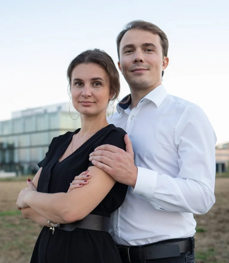 Ein Mann und eine Frau stehen draußen, der Mann hat seinen Arm um die Schulter der Frau. Es ist bewölkt, im Hintergrund sind moderne Gebäude zu sehen.