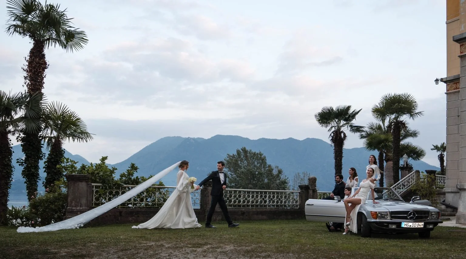 Hochzeit im Freien mit einer Braut in Weiß, einem Bräutigam in schwarz, und Gästen, die an einem schwarzen Mercedes-Cabrio posieren, umgeben von Palmen mit Berg im Hintergrund.