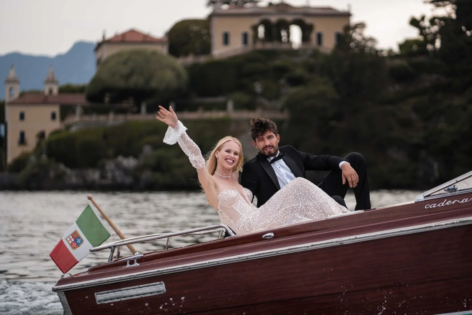 Ein lachendes Paar in formeller Kleidung, wahrscheinlich Hochzeitskleidung, fährt auf einem Boot auf Wasser, mit einem italienischen Flagge am Bug, während im Hintergrund eine Villa auf einem Hügel steht.