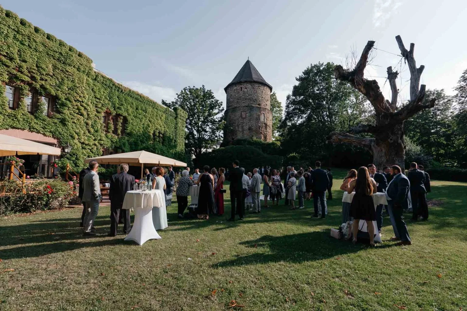 Hochzeit im Garten mit alten Turm und großen Baum, Gäste in formeller Kleidung, Stehtische, Sonnenschein, grüne Umgebung.